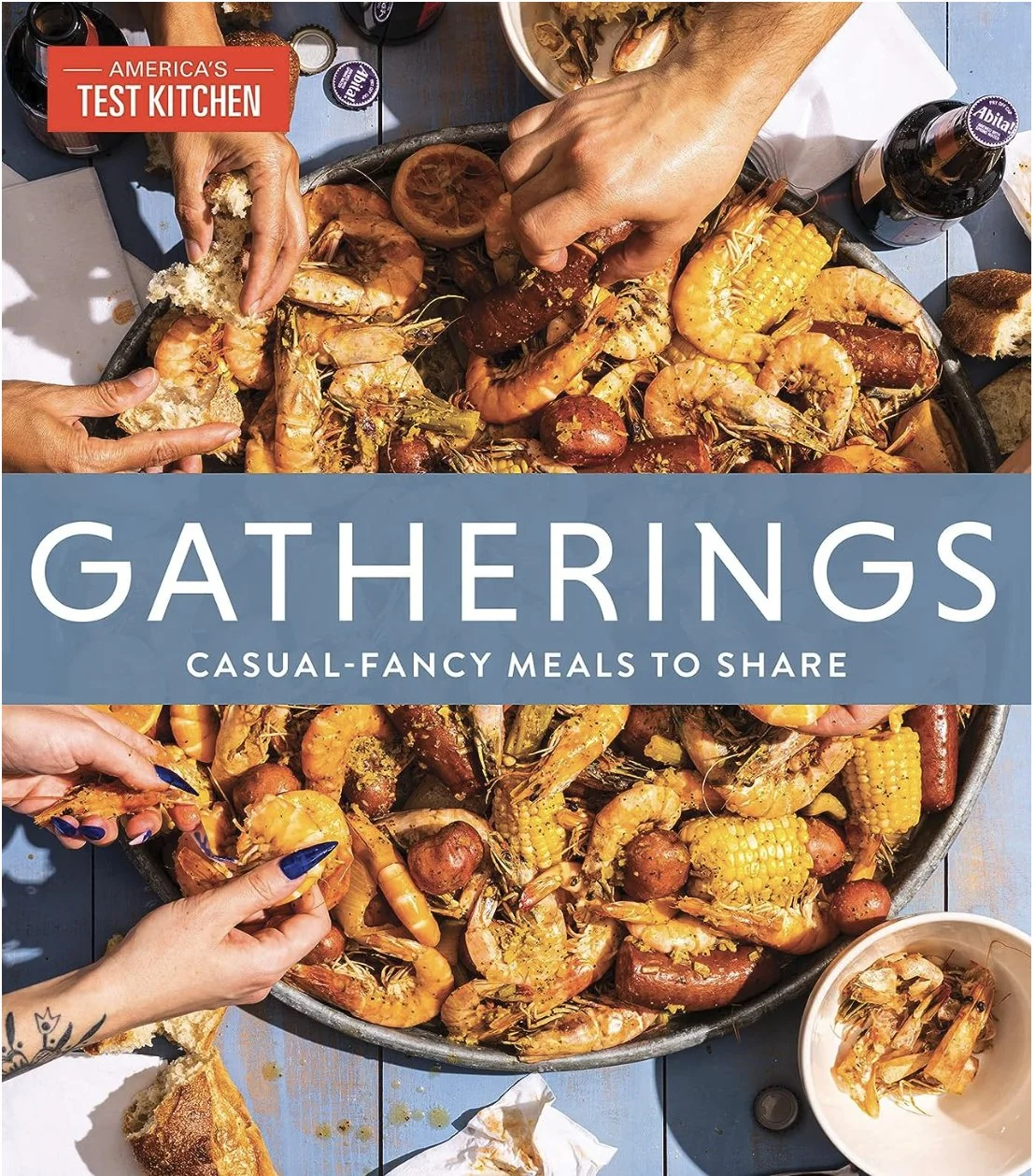 Cover of the book, Gatherings by Americas Test Kitchen. A large platter of cooked seafood including shrimp, lobster, and corn on the cob, with hands reaching in to grab food, on a blue wooden table with condiments and bread nearby. 