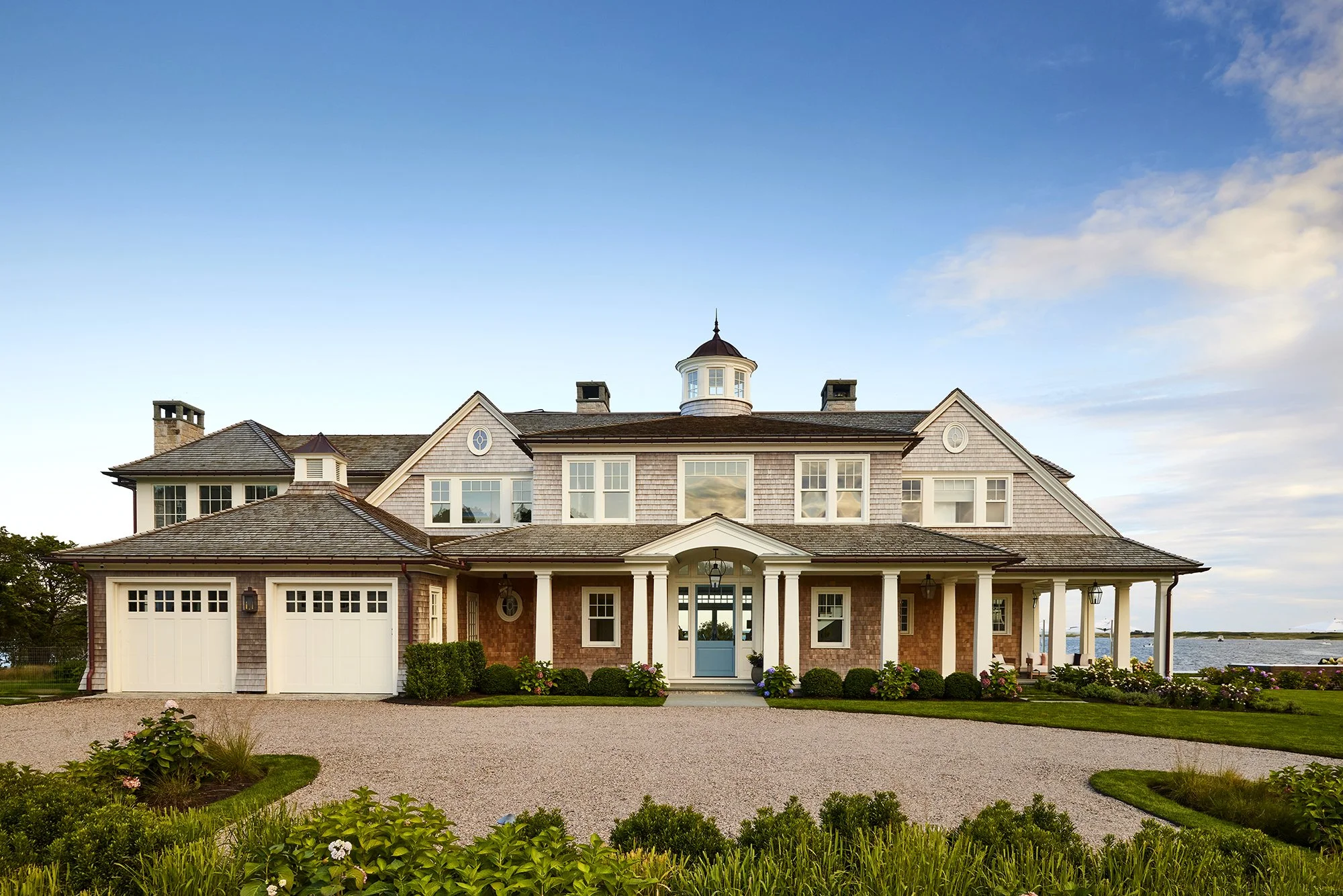Large, elegant house with a brick and wood exterior, multiple windows, a porch with white columns, and a gravel driveway, overlooking a body of water under a blue sky with some clouds.