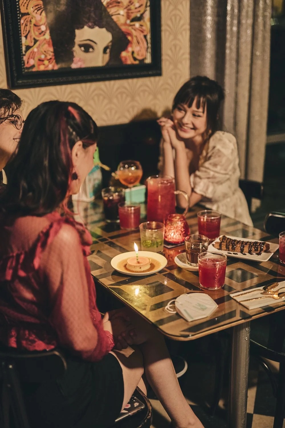 A group of four women celebrating a birthday at a restaurant, with one woman in the foreground having a birthday cake with a sparkler candle. There are various drinks, a plate with a sushi roll, and a lit candle on the table. One woman is smiling and