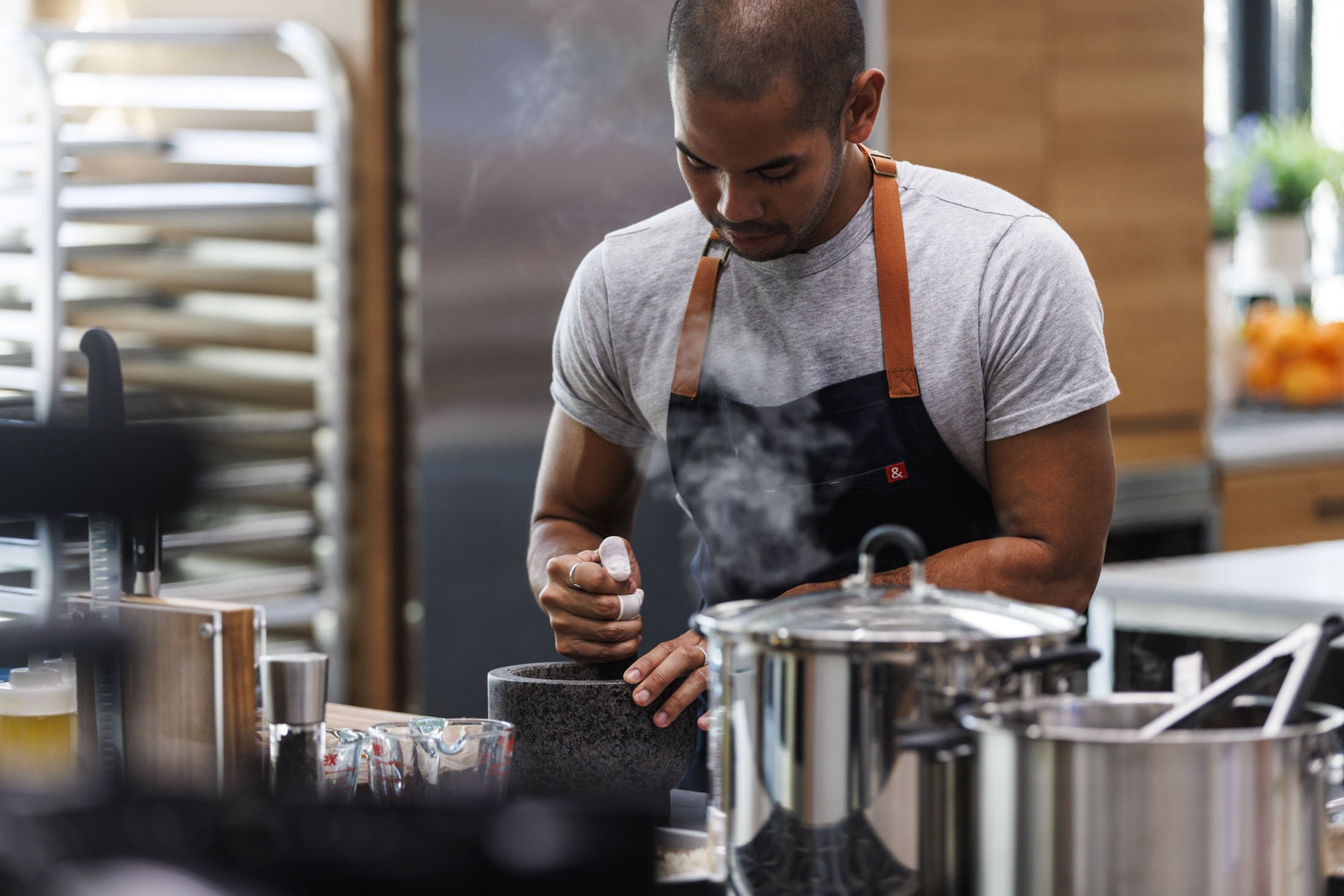 A man wearing a gray t-shirt and apron grinding spices in a kitchen. There are stainless steel pots and kitchen utensils on the counter, with a rack of baking trays and a window with plants in the background.