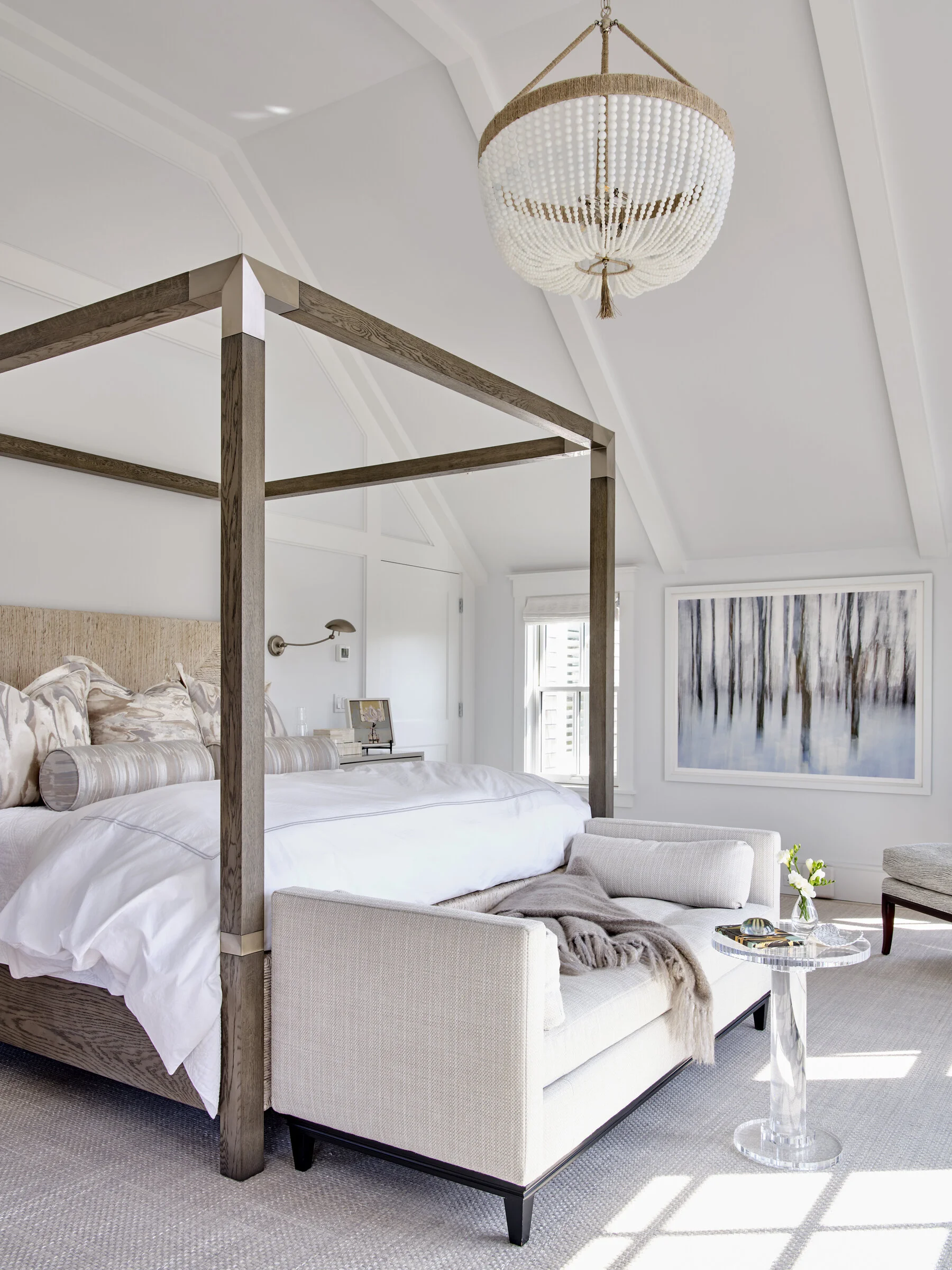 A bright bedroom with a four-poster bed, white bedding, a cream sofa with a throw, a small glass side table with flowers, wall art, and a beaded chandelier hanging from the ceiling. Professional architectural photograph