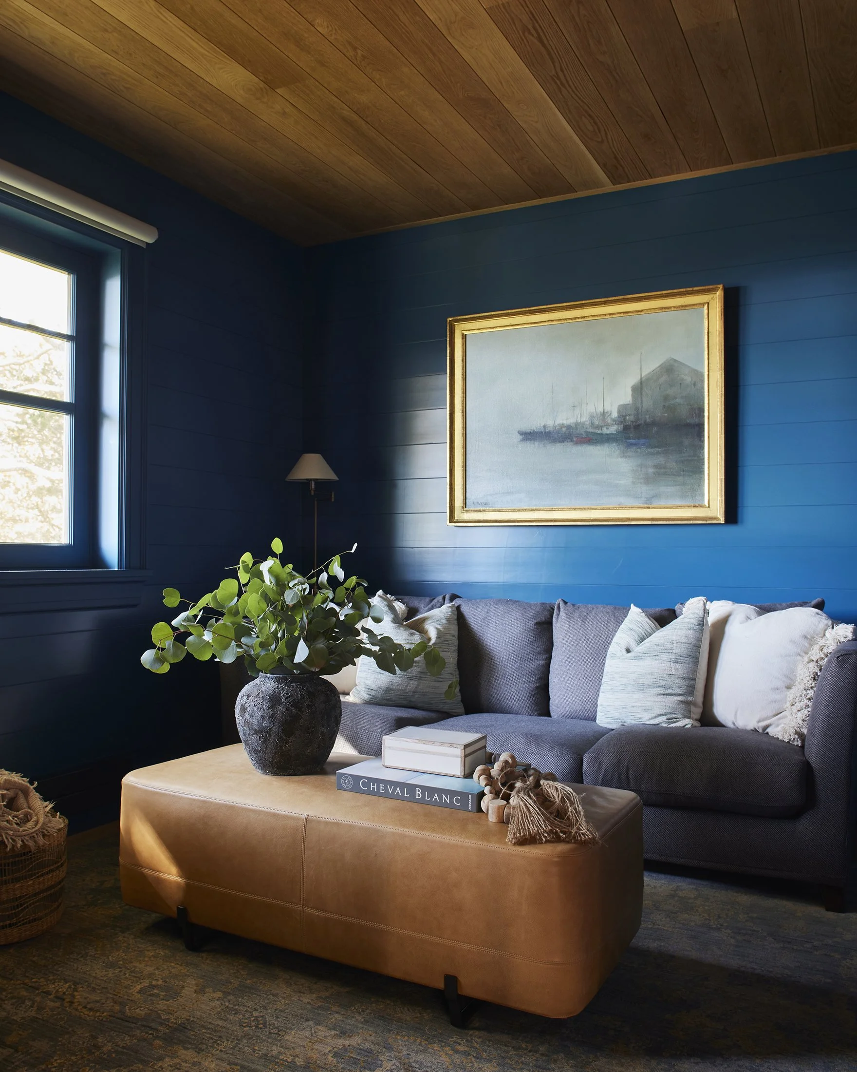 Living room with dark blue walls, wooden ceiling, and a large window. A gray sectional sofa with white and green pillows, a tan ottoman with a black vase of green leaves, books, and decorative items, and a framed painting of a harbor scene on the wal