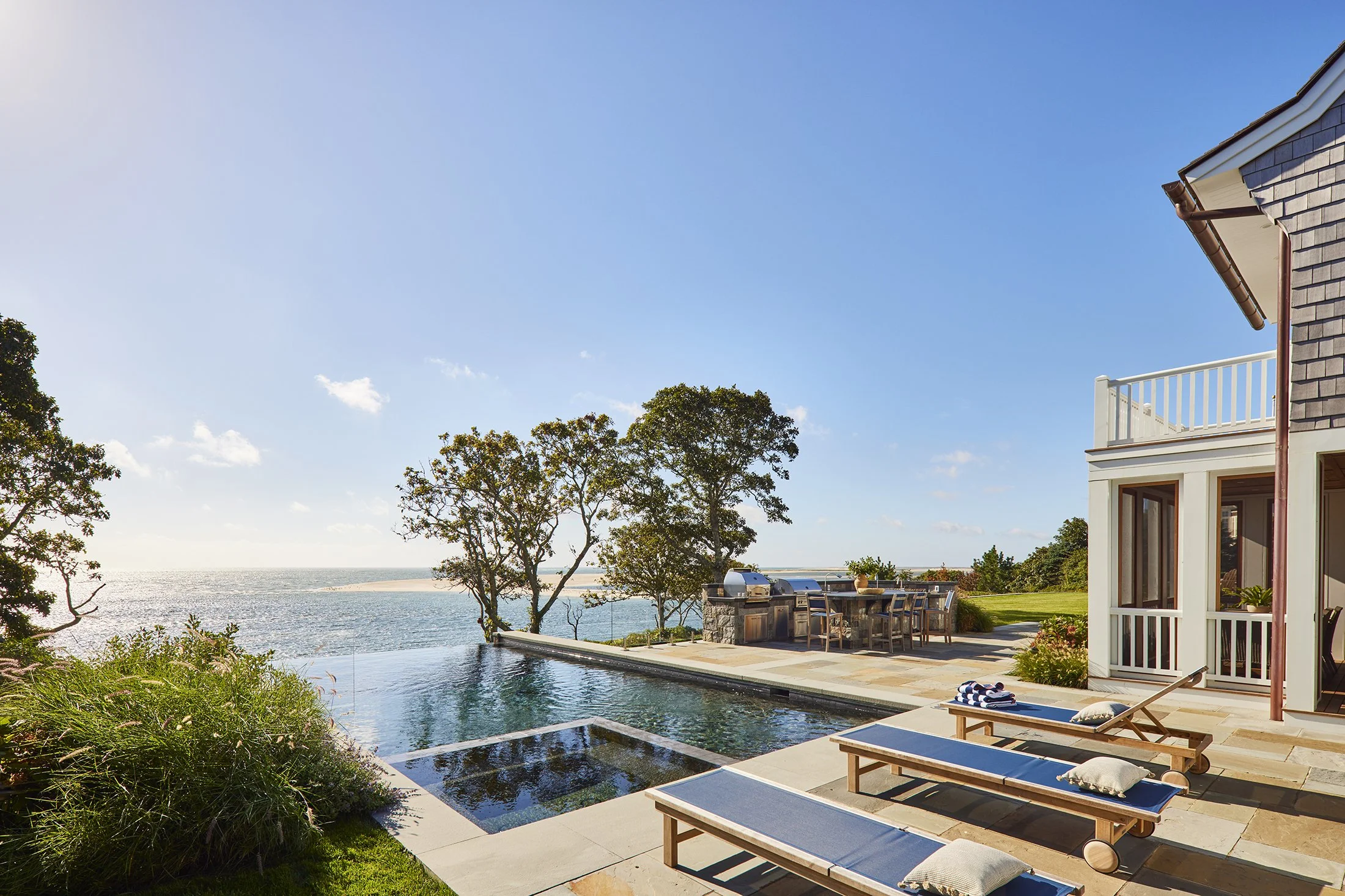 Luxury house with an infinity swimming pool overlooking a body of water, outdoor dining area, lounge chairs and trees under a blue sky. Professional architectural photograph of a coastal Cape Cod swimming pool.