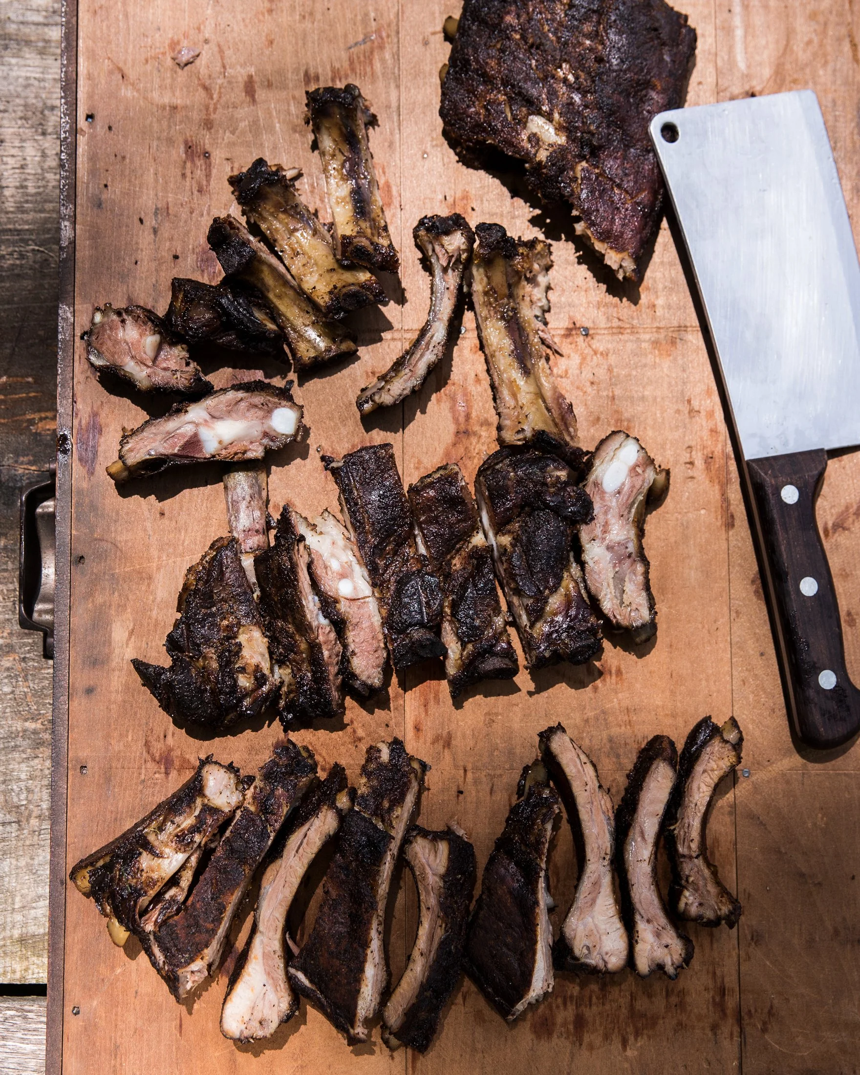 Smoked beef ribs arranged on a wooden cutting board with a meat cleaver nearby.