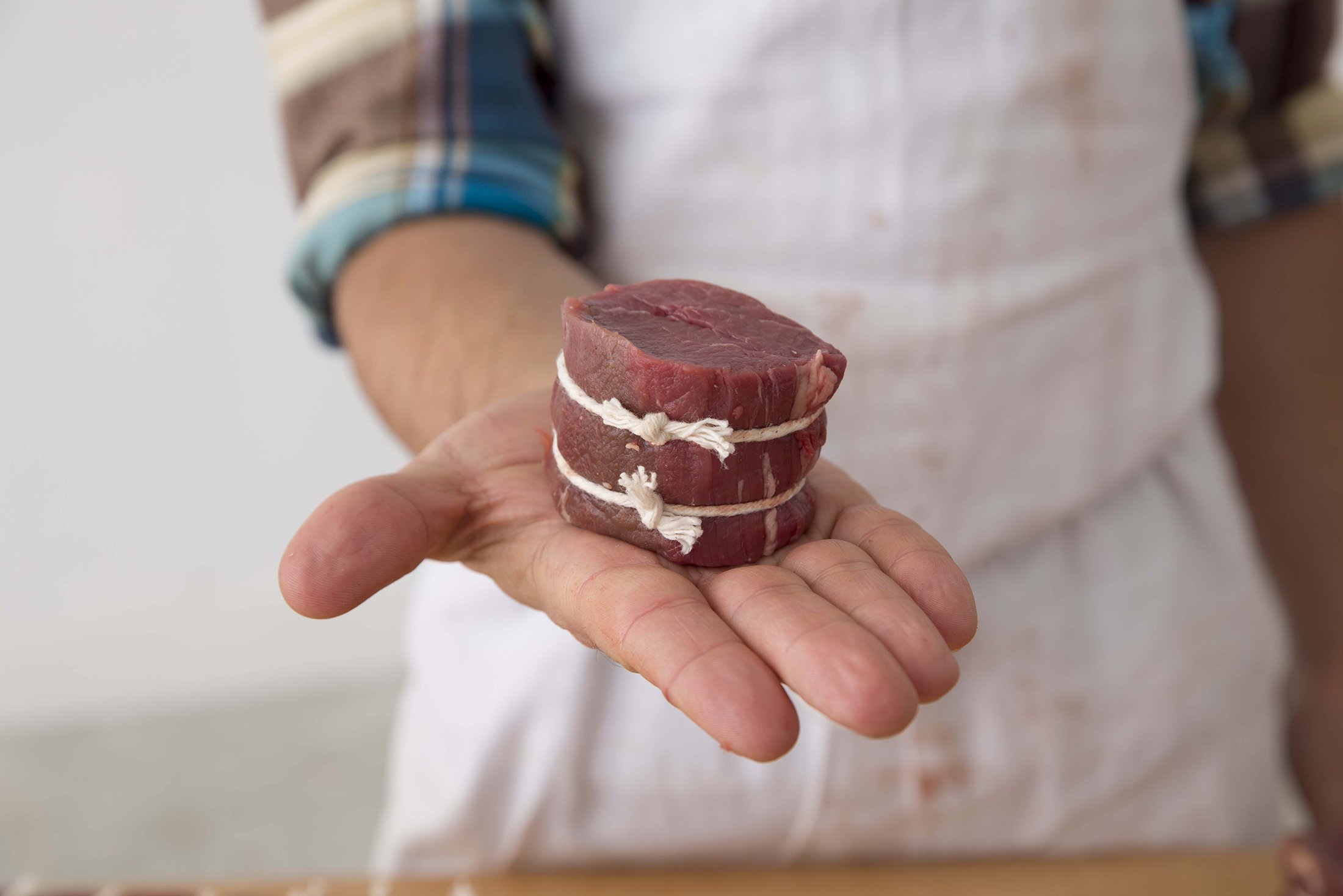 Butcher holding two cuts of raw beef steak tied with white string in a kitchen setting.