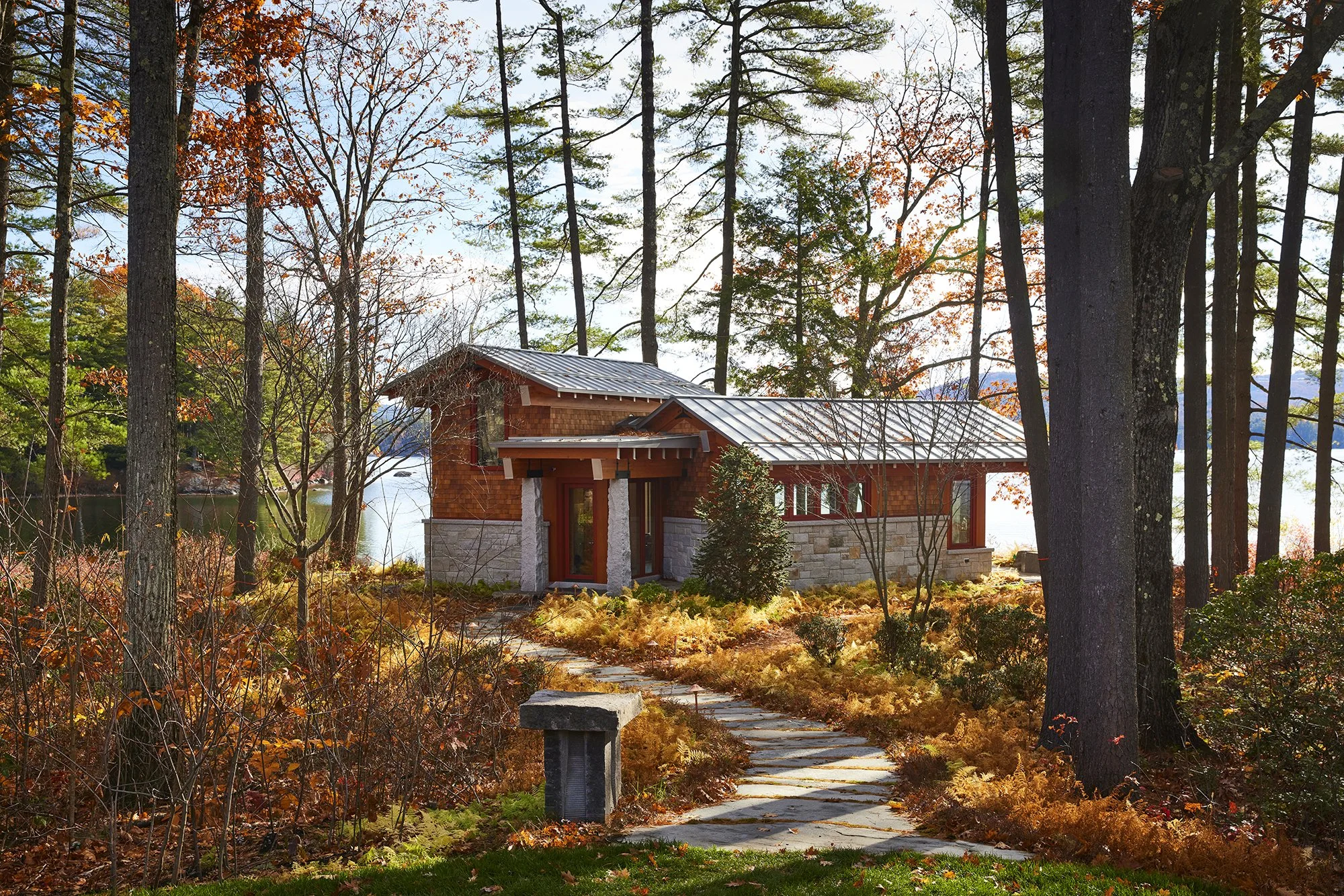 A small modern house with wooden and stone exterior walls located in a wooded area near a lake, surrounded by autumn-colored trees and a winding stone pathway leading to the entrance. Professional architectural photograph of a lakeside guest house.