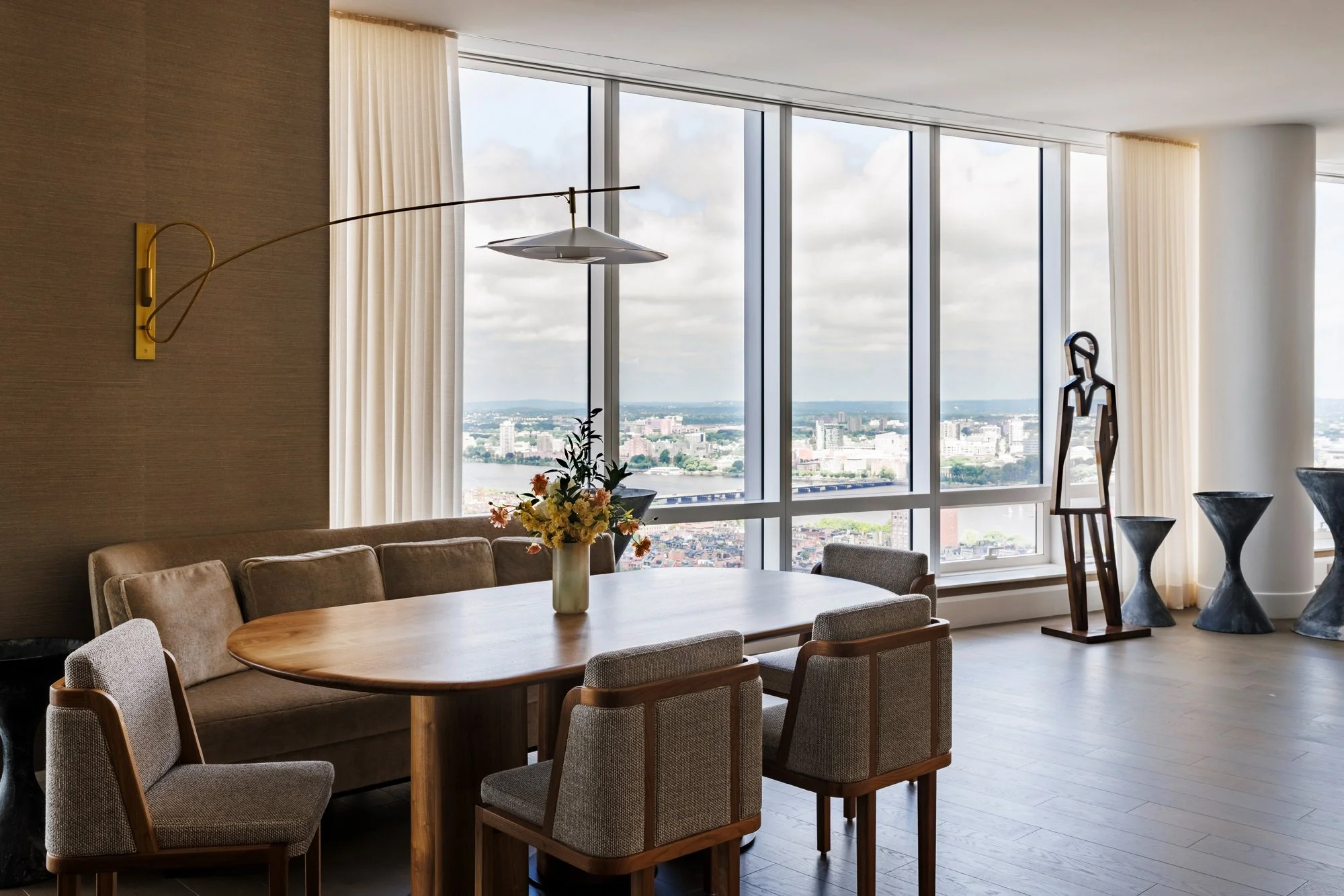 Modern dining room with a large oval wooden table, six upholstered chairs, a beige sofa, a flower vase, and large floor-to-ceiling windows offering a cityscape view. Art sculptures and tall stone-like tables are in the background.