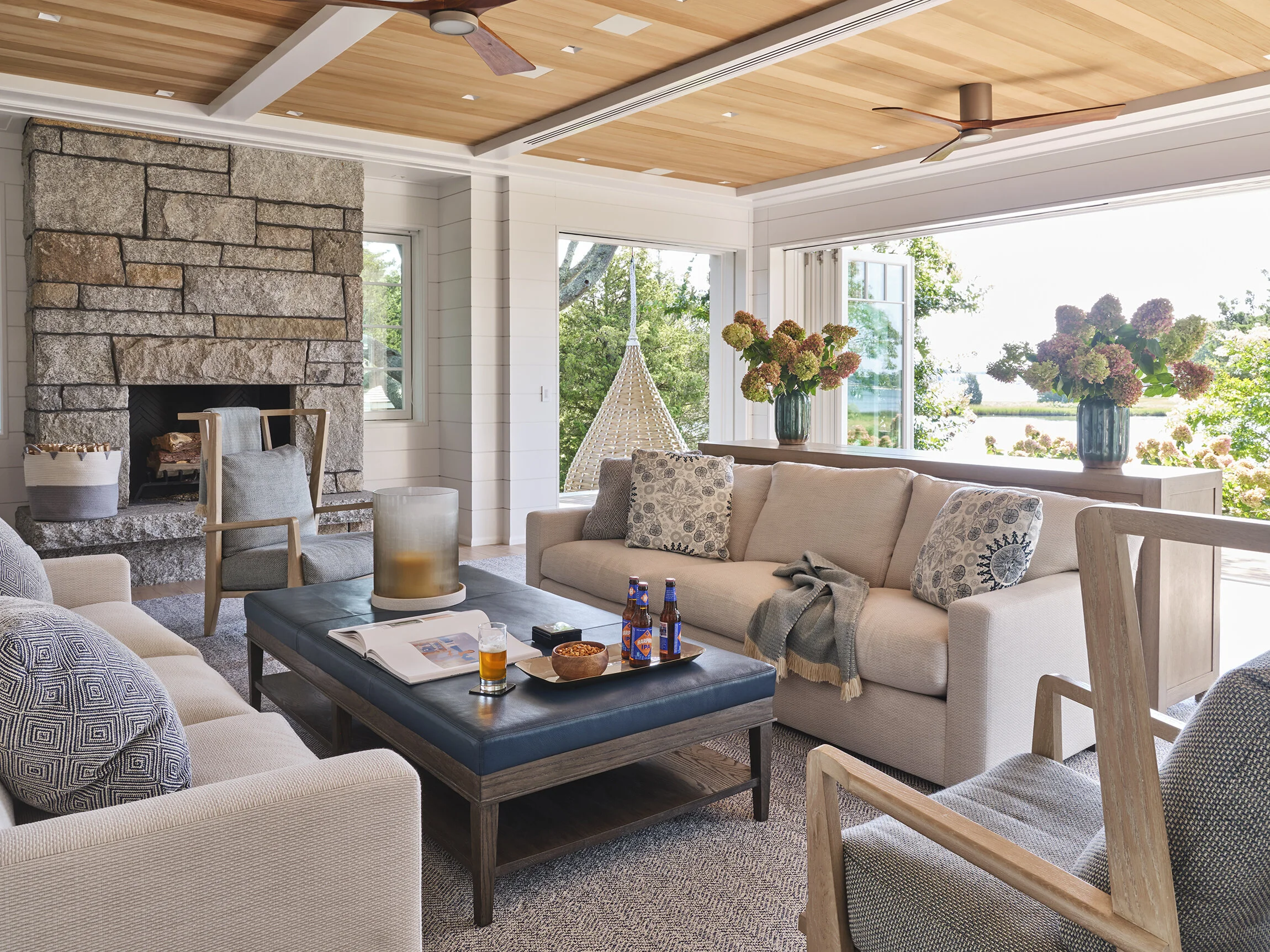 Living room with white sofa, armchairs, stone fireplace, wooden ceiling with ceiling fans, large windows with view of greenery, and vases with pink flowers.