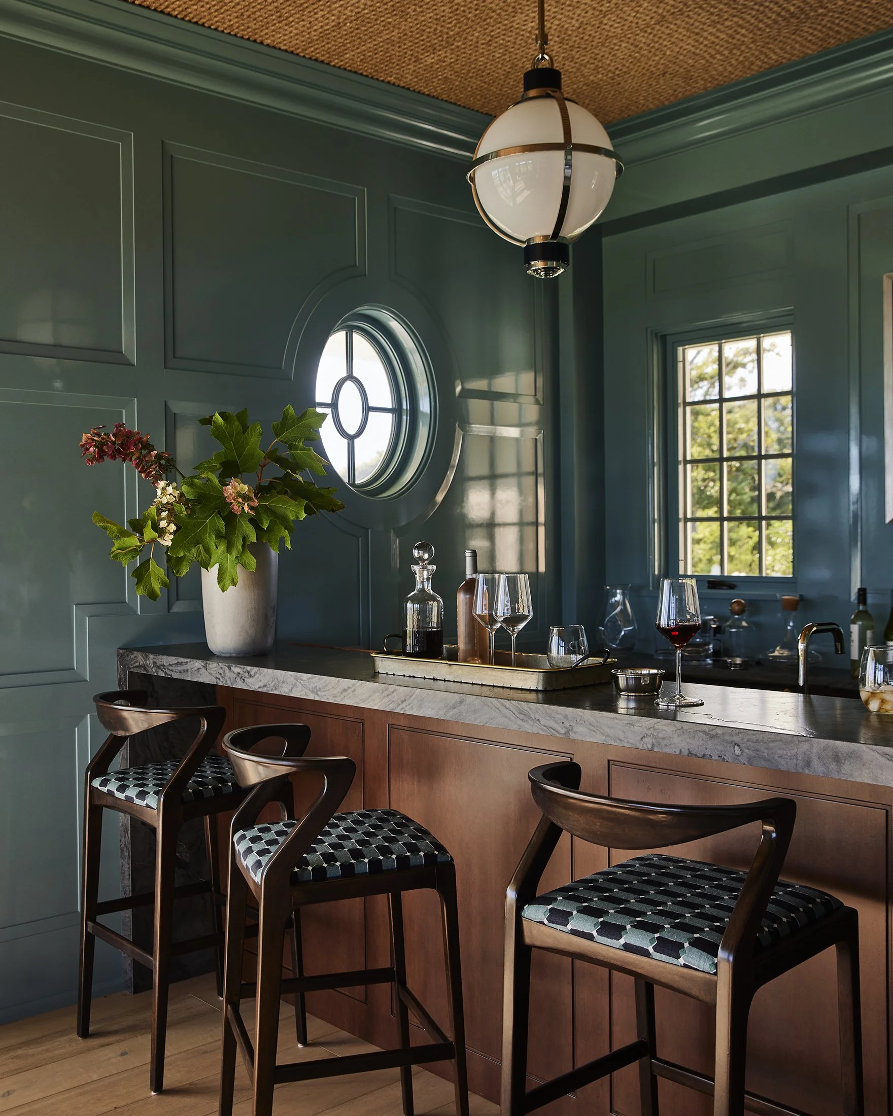 Interior of a home bar with green walls, a round window, dark wood bar stools with patterned cushions, a marble countertop with wine glasses, bottles, and a large vase with greenery, a window showing greenery outside, and a ceiling lamp.