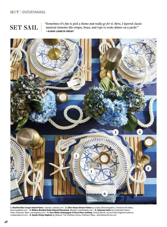 A nautical-themed table setting with blue, white, and gold accents, featuring plates, glasses, flowers, and decorative objects.