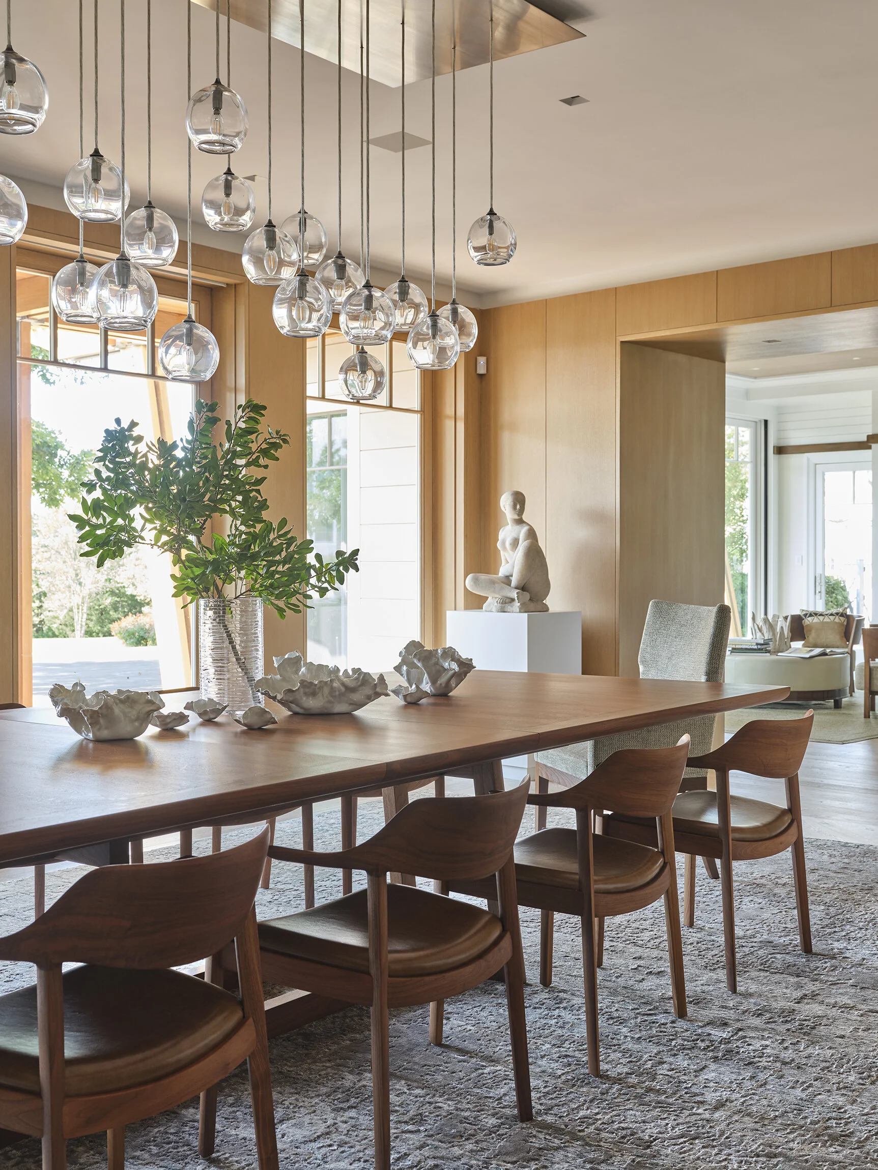 Modern dining room with wooden table, six chairs, glass pendant lights, decorative ceramics, a large green plant, and a sculpture of a seated woman.