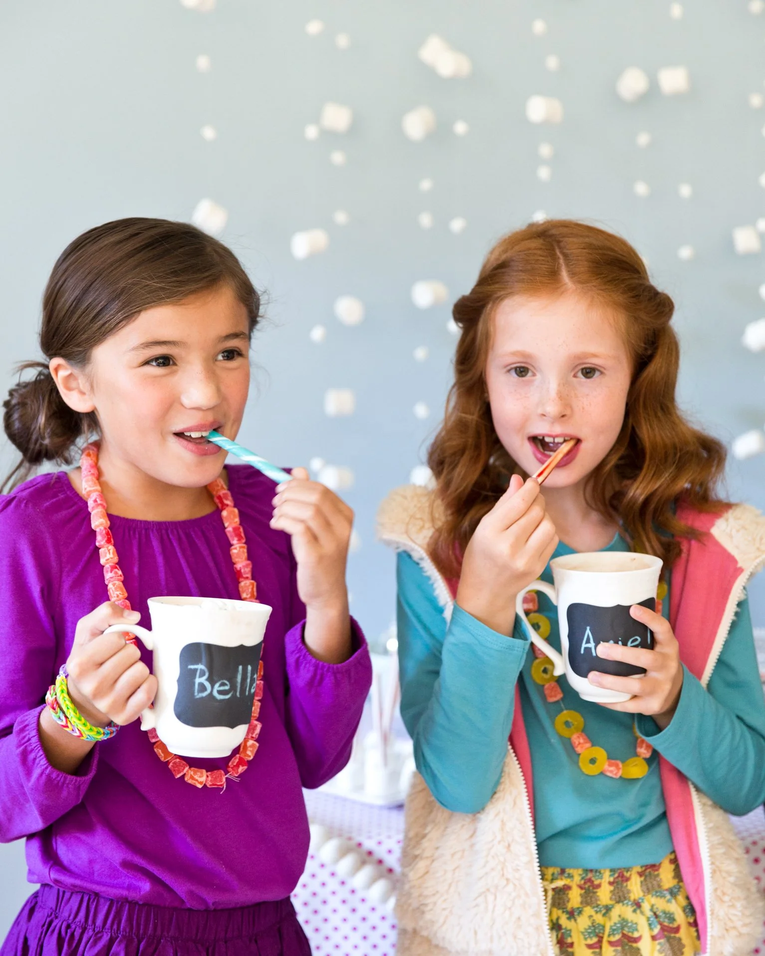 Two young girls celebration with cups and party hats, one with dark hair in a purple dress and the other with red hair in a teal shirt and colorful skirt, surrounded by confetti.