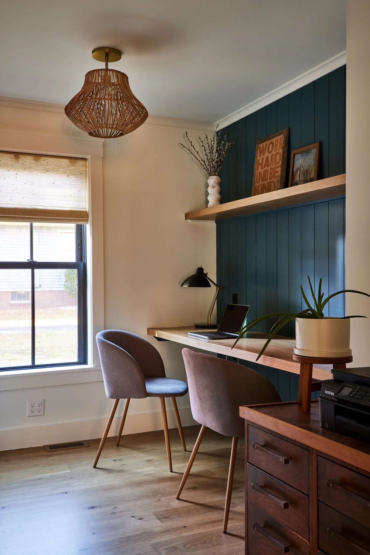 Home office with a window, a desk against a blue wall, two pink and purple chairs, a black desk lamp, a potted plant, and decorative items on a shelf.