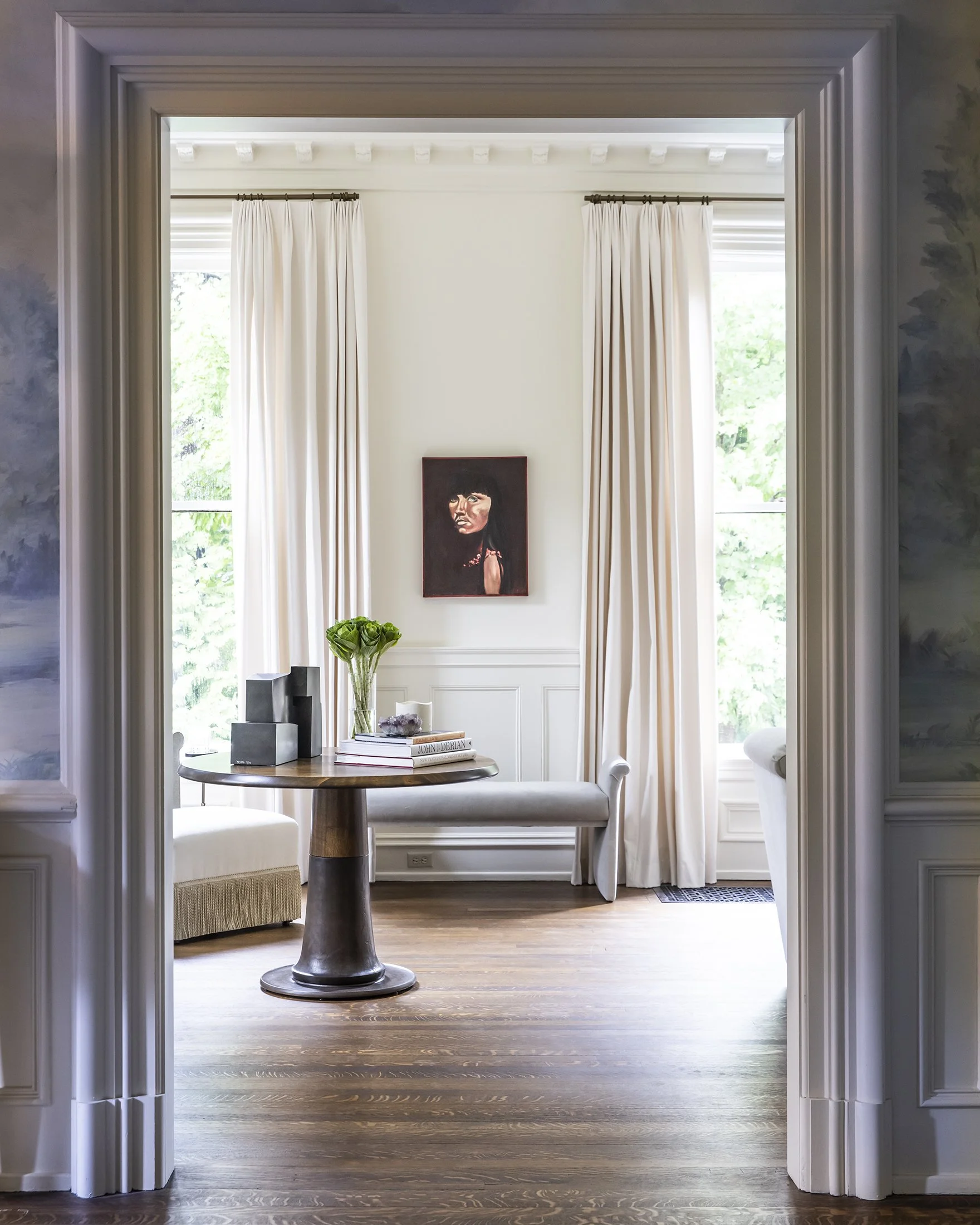 View of a bright room with large windows with white curtains, a round wooden table, a bench, books, a plant, and a portrait painting on the wall.