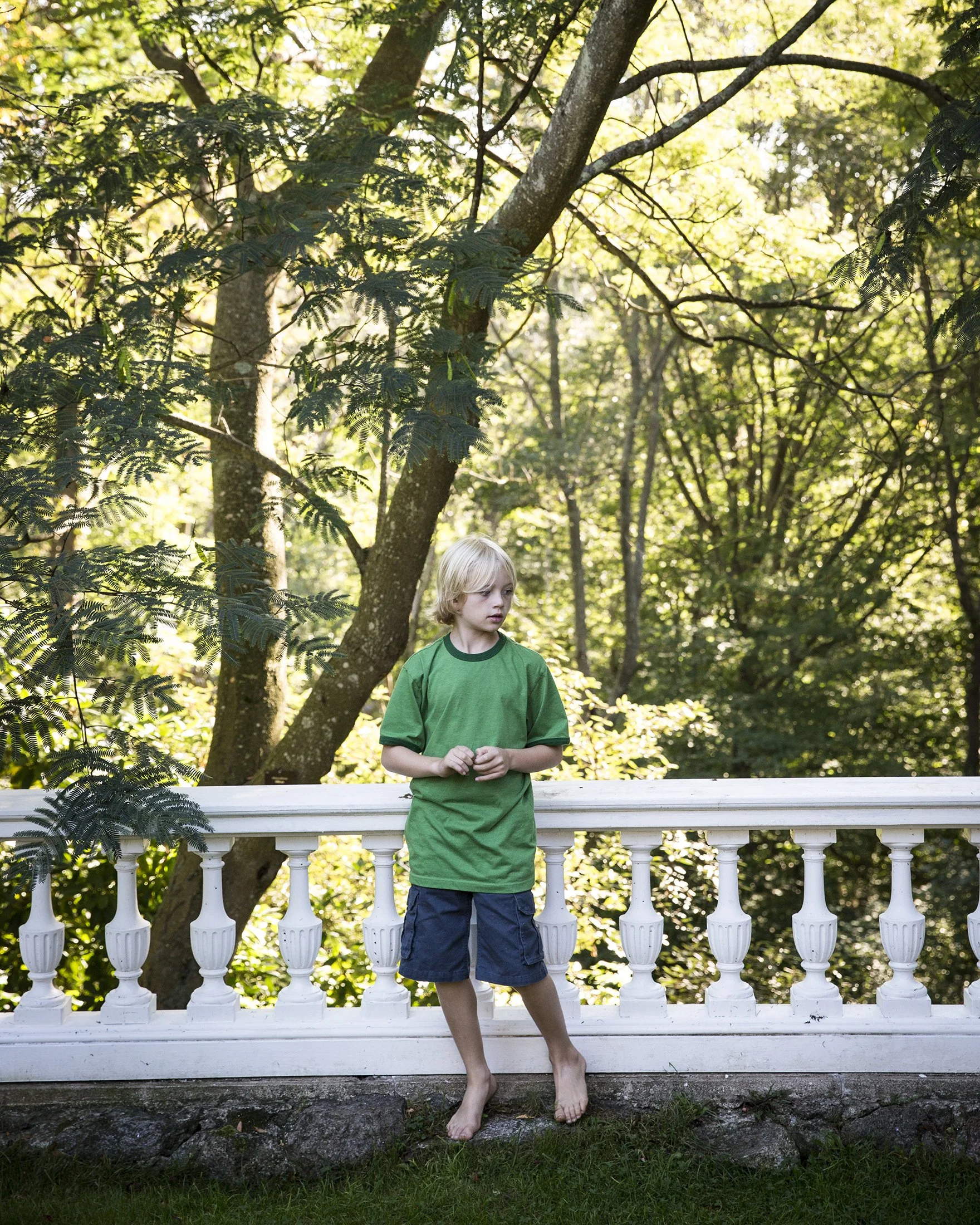 A young boy with blonde hair, wearing a green t-shirt and navy shorts, standing barefoot on the grass next to a white decorative railing, looking to the side amidst a lush, sunlit forest.