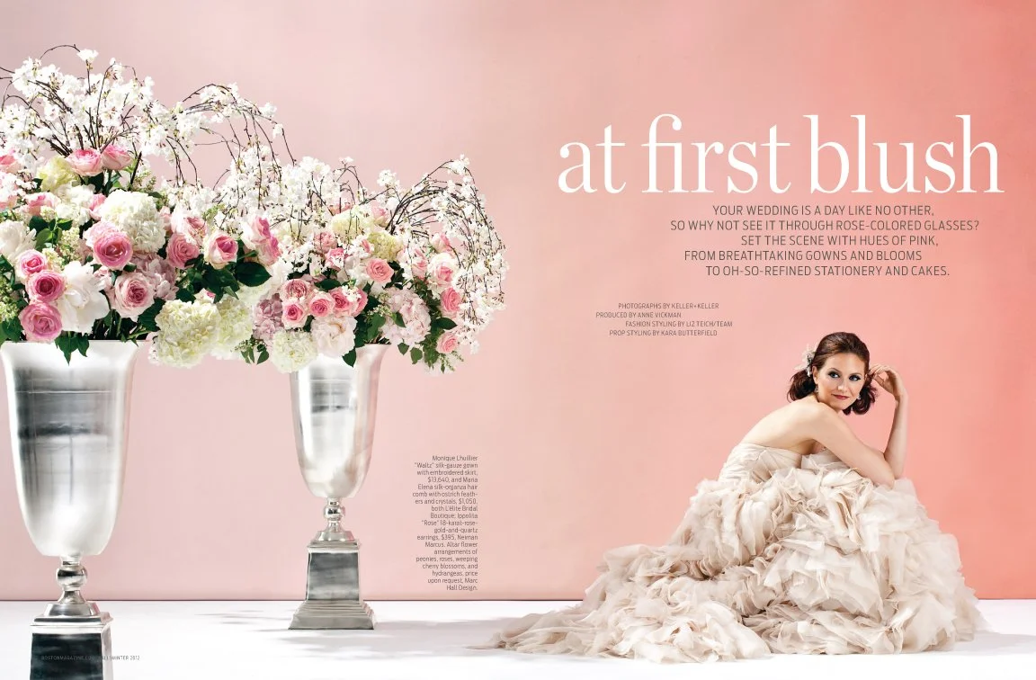 A woman in a flowing, ruffled wedding gown sitting on the floor with her head resting on her hand, next to three tall silver vases filled with pink and white flowers against a pink background.