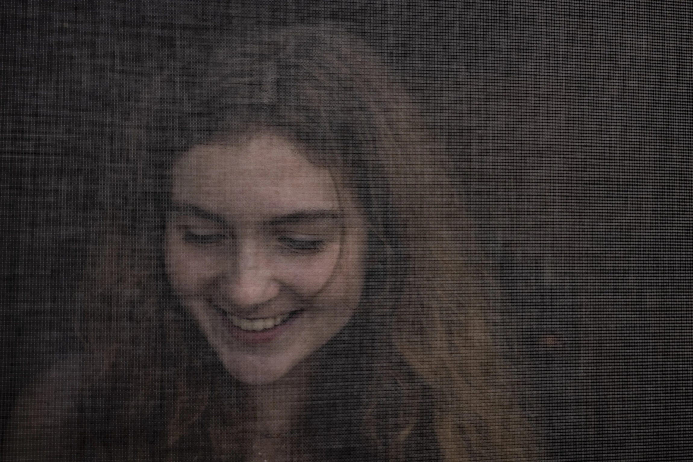 A young woman with long, wavy hair smiling through a screen or mesh.