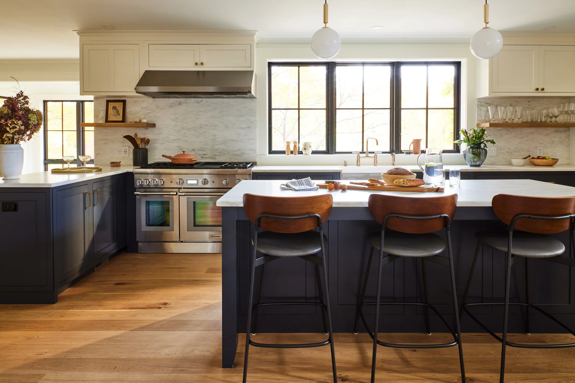 Modern kitchen with white countertops, black cabinets, a large window, and a kitchen island with three wooden and metal chairs, featuring baked goods, a water pitcher, and glasses on the island.
