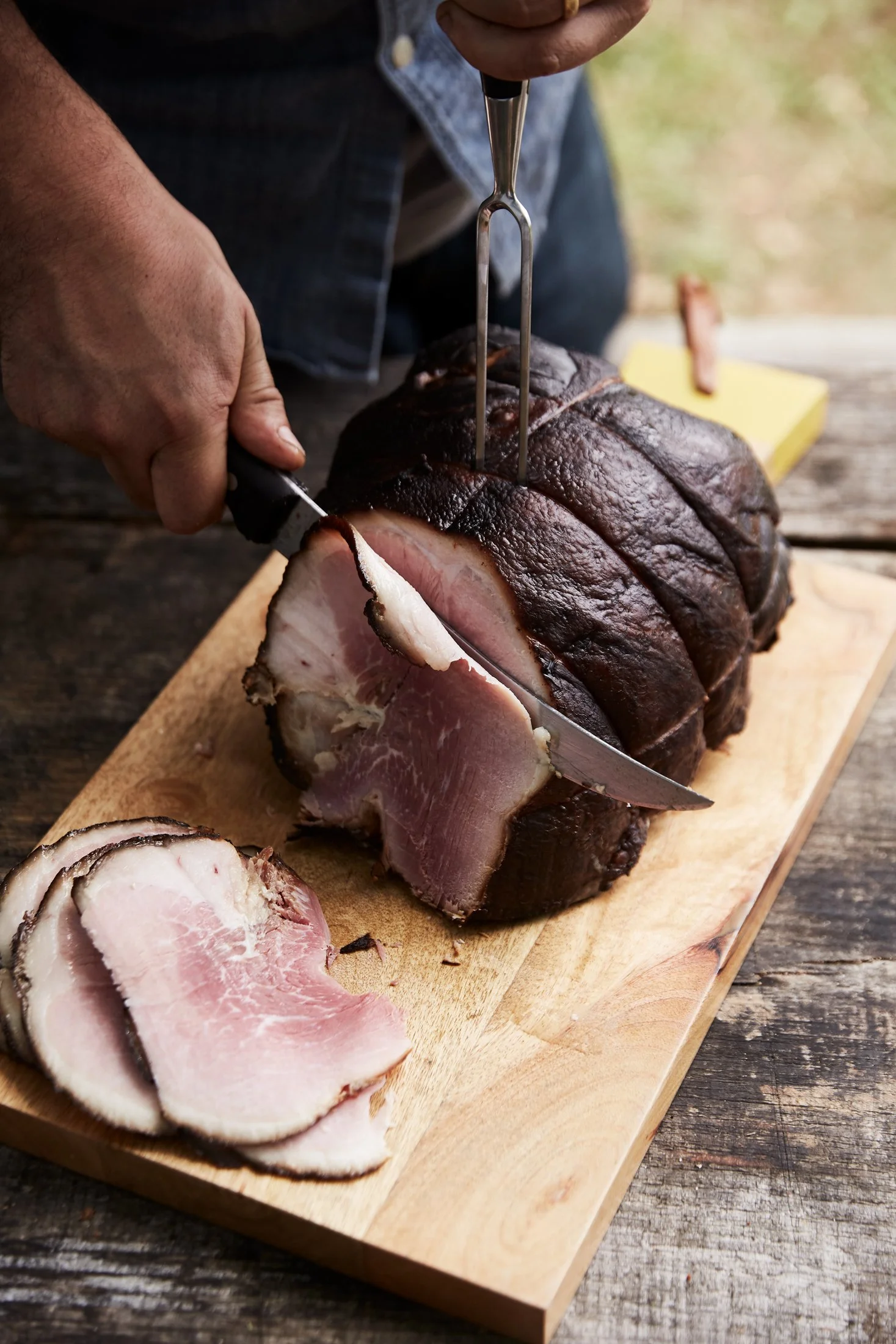 A person is slicing smoked or cured meat, possibly ham or pork, on a wooden cutting board outdoors, using a sharp knife and holding the meat in place with a meat fork.