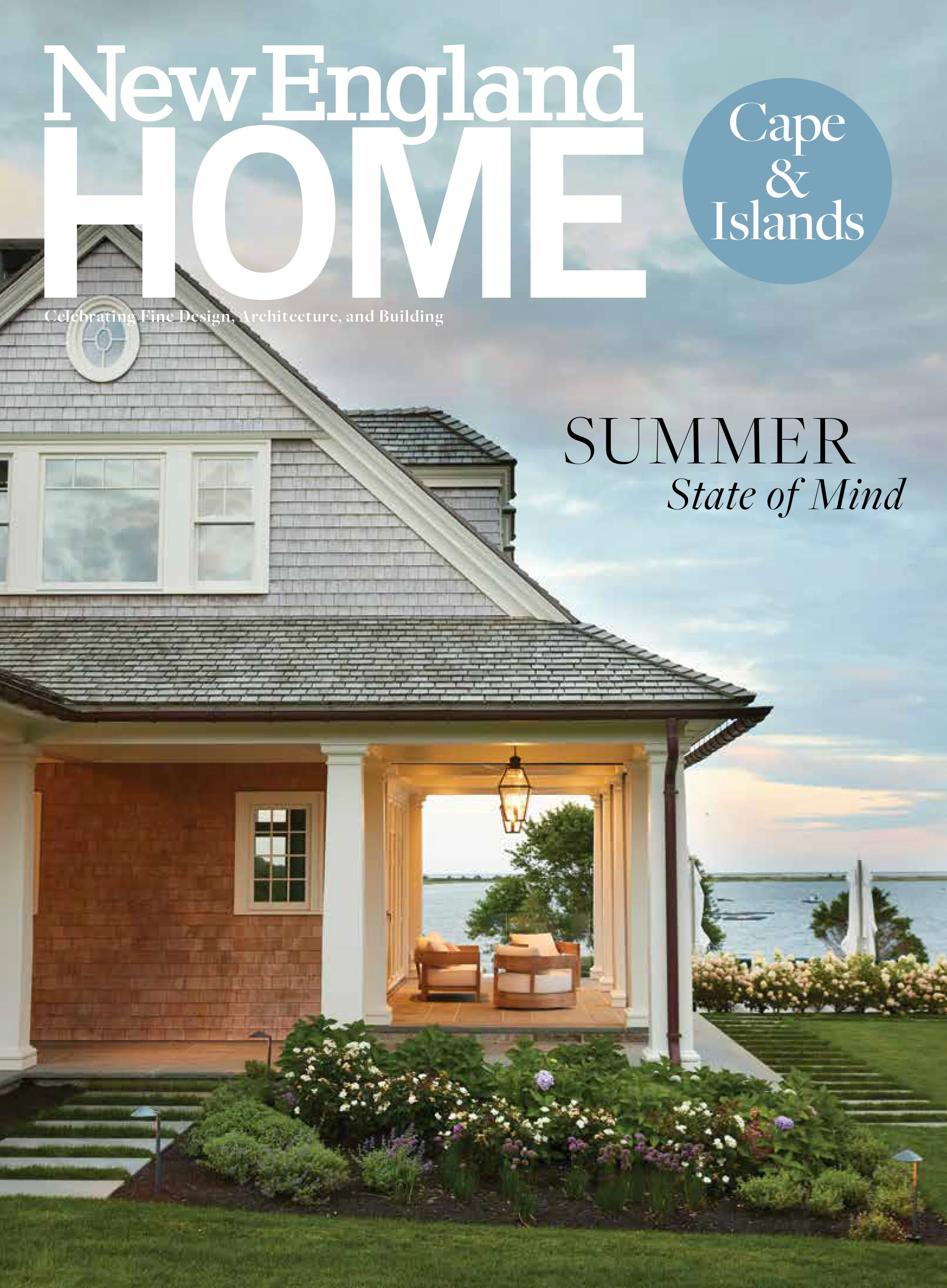 Cover of 'New England Home' magazine featuring a house with a porch overlooking a body of water, floral garden in the foreground, and a partly cloudy sky.