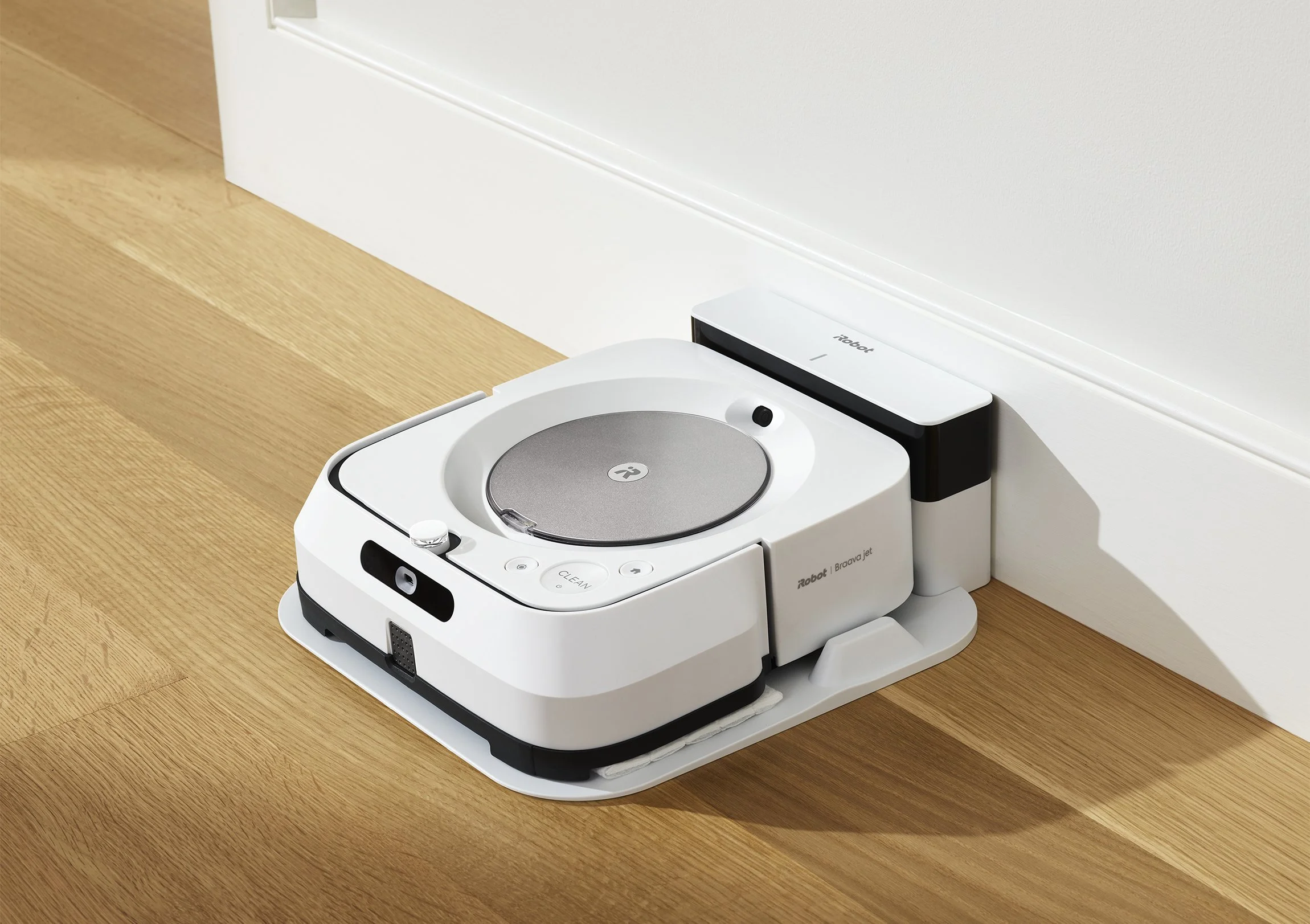 A robotic vacuum cleaner docked against a white wall on a hardwood floor. Photographed by Joseph Keller for iRobot