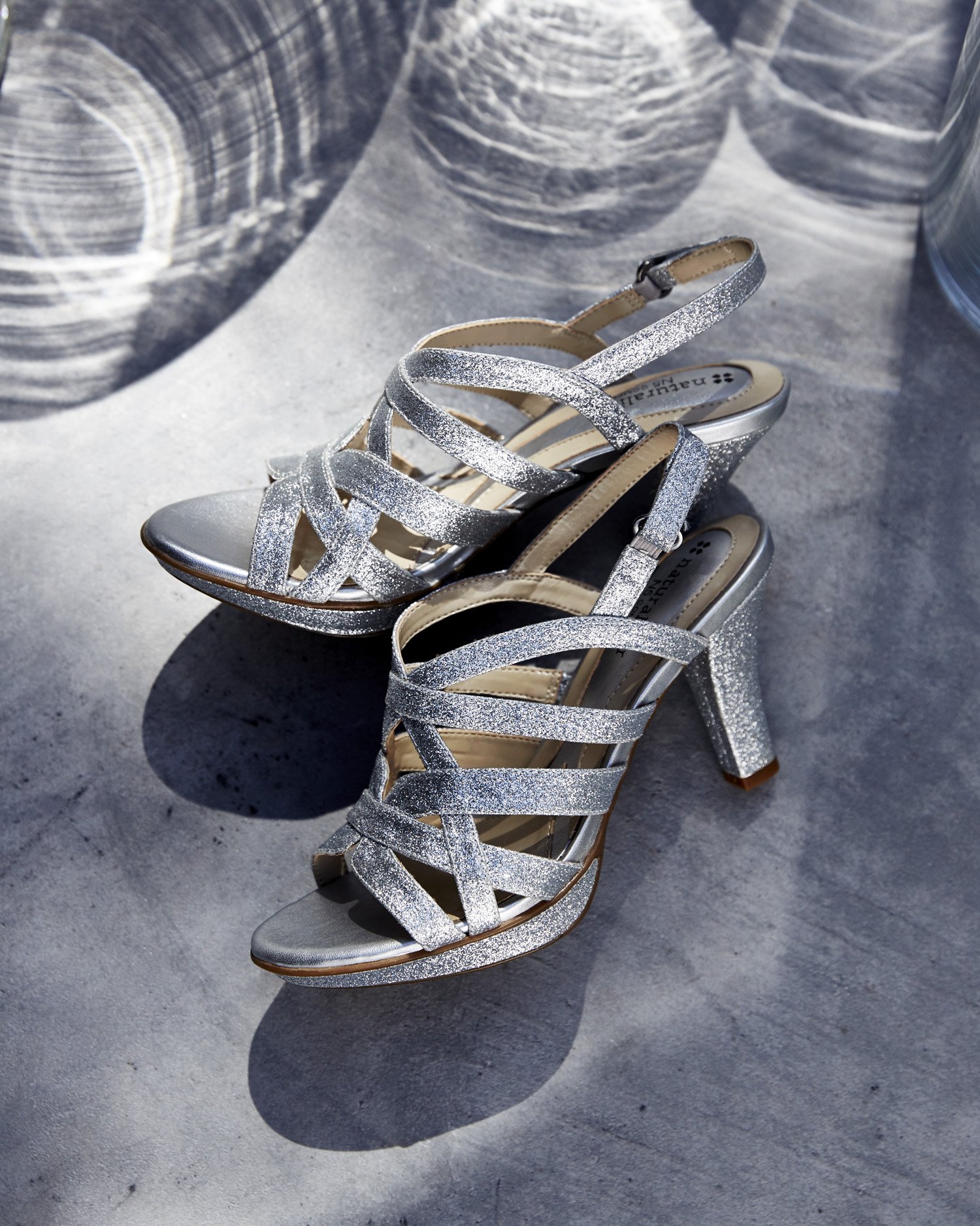 Silver glittery high-heeled sandals with multiple straps, placed on a concrete surface with shadows.