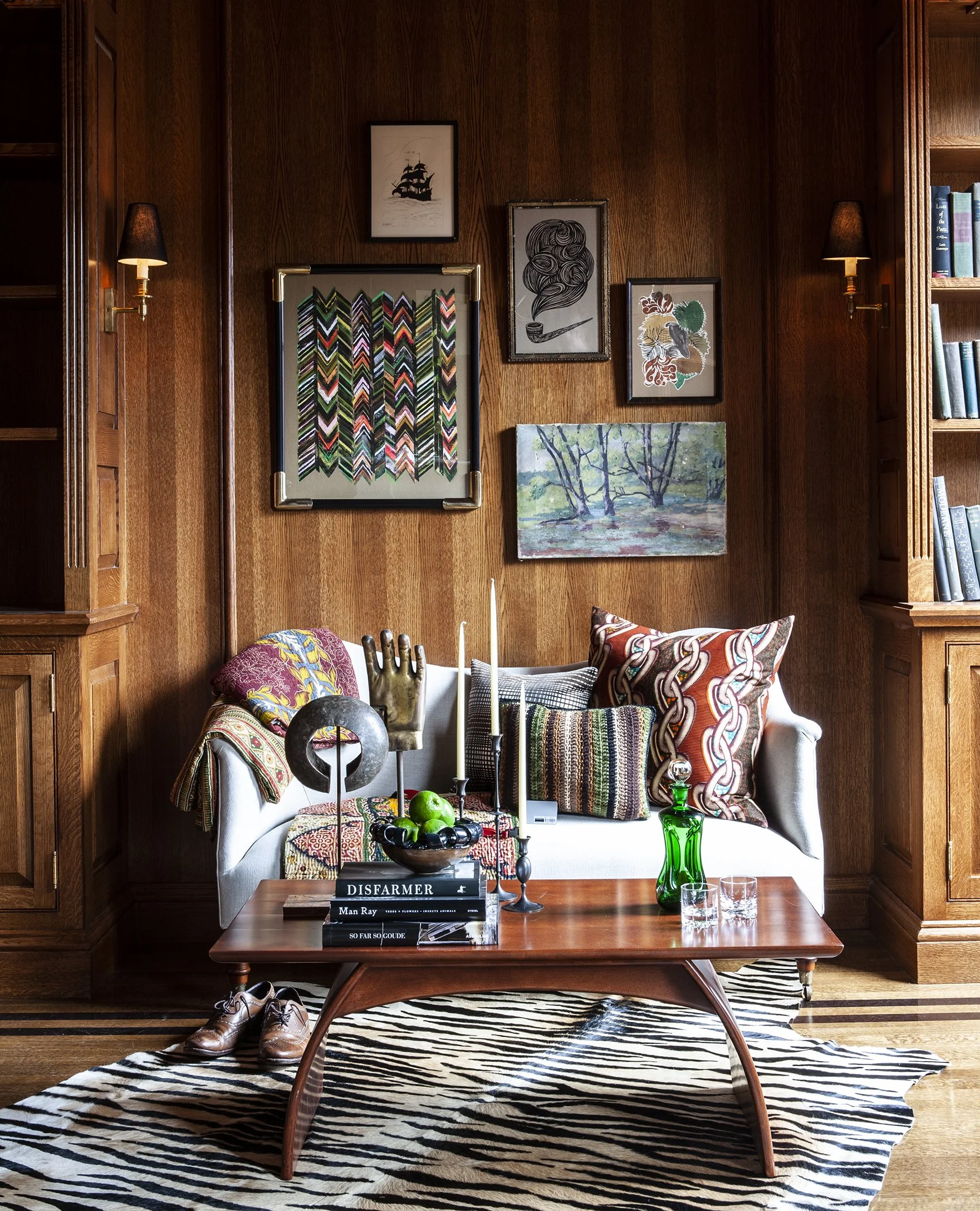 A living room corner with wood-paneled walls, a white sofa with colorful patterned pillows, a wooden coffee table with books, candles, and a green glass bottle, a zebra print rug, and framed artwork on the wall.