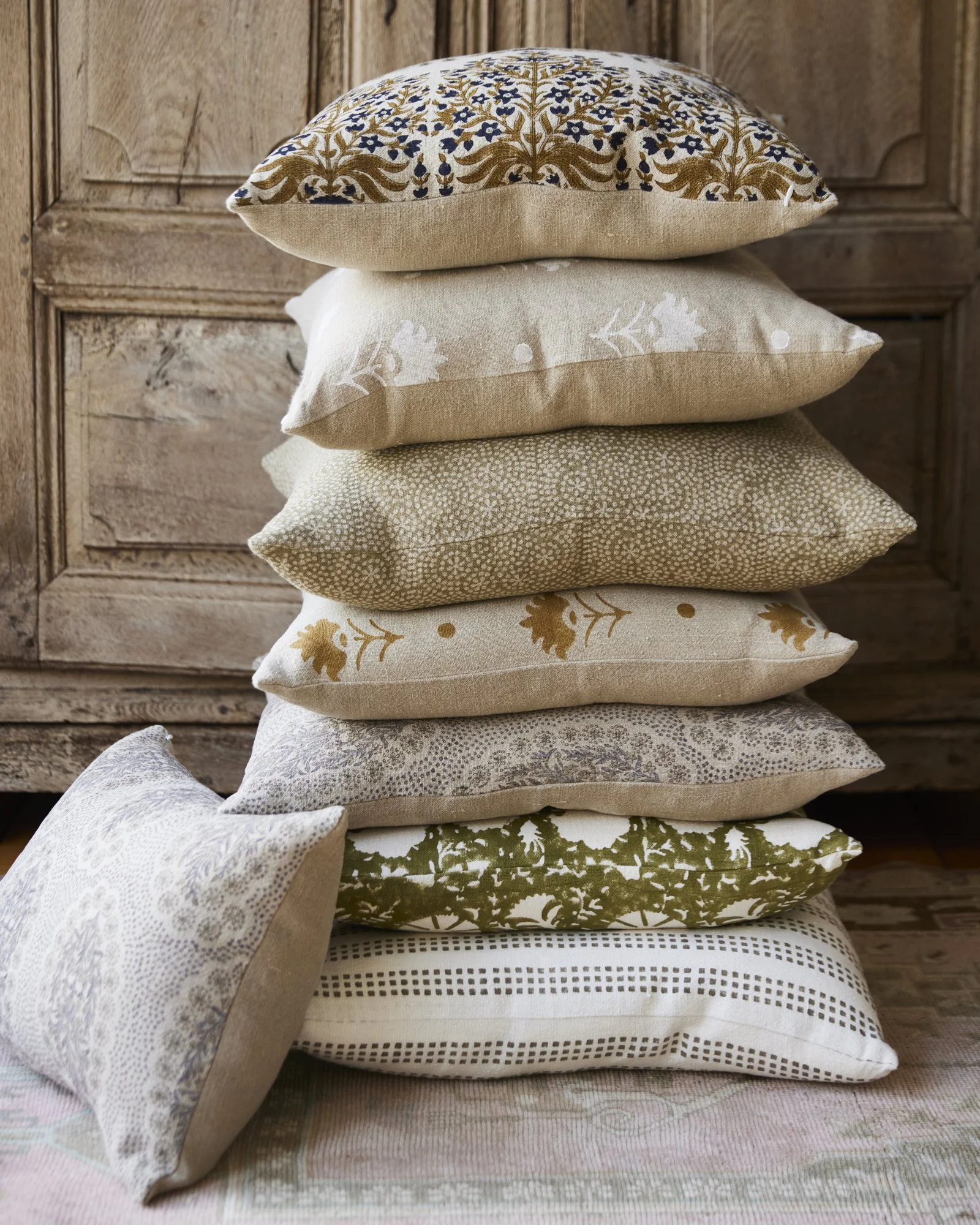 Stack of seven decorative pillows with various neutral and earth tone patterns, placed on a floral-patterned rug in front of wooden furniture. Photographed for Duxbury Supply Co. based in Duxbury MA.