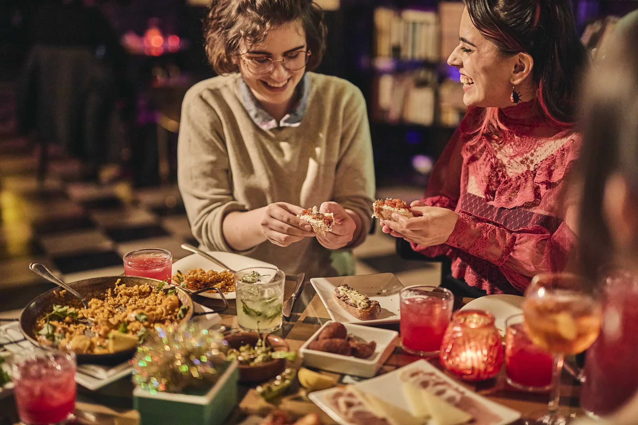 Two women laughing and sharing food at a dinner table with various dishes, drinks, and candles, in a warmly lit setting. Boston hospitality photographer.