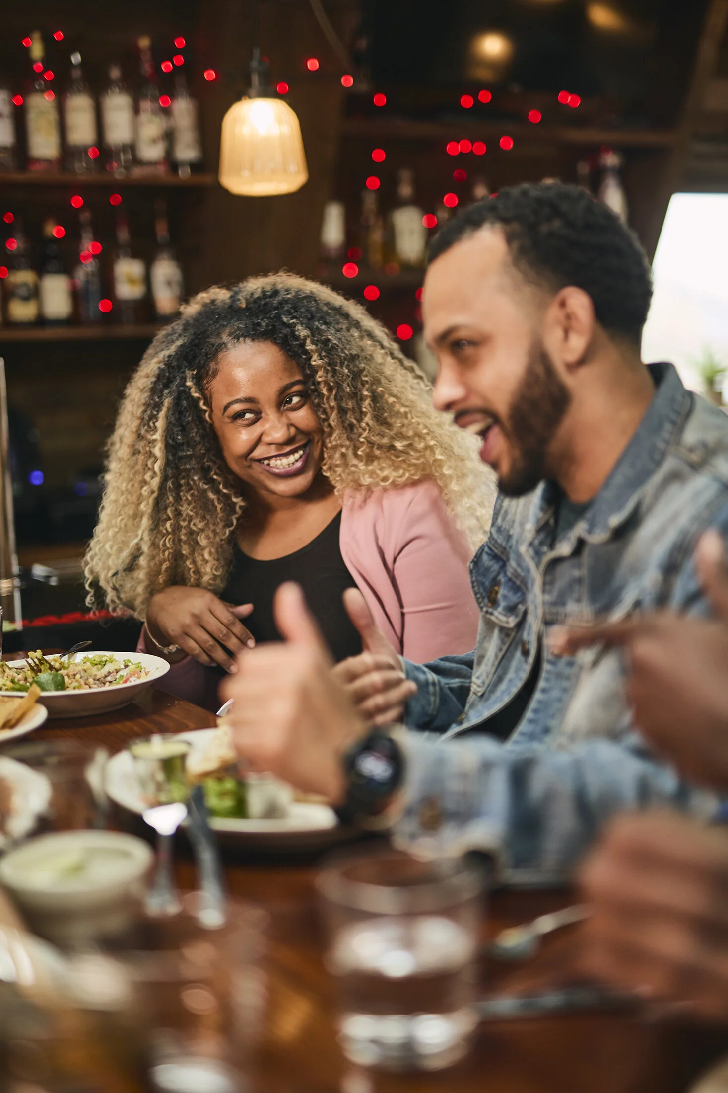 A group of friends enjoying a lively conversation at a restaurant table, with food and drinks, celebrating and smiling.