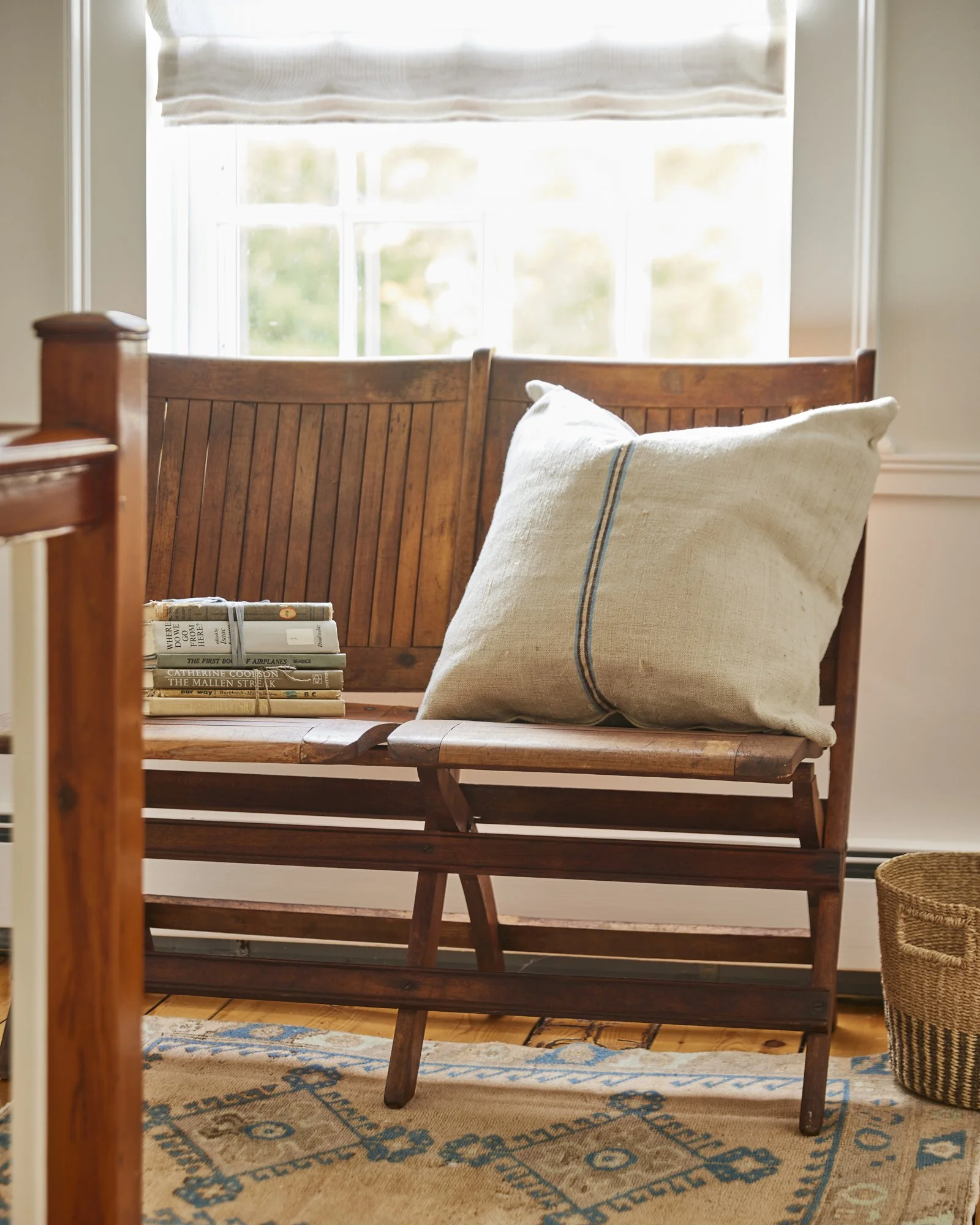 Wooden bench with beige cushion and a stack of books, placed in front of a window with natural light. Photographed for Duxbury Supply Co. based in Duxbury MA.