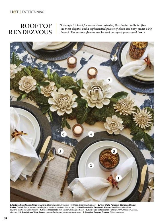 A beautifully decorated outdoor table setting on a rooftop with a floral centerpiece, white plates, gold utensils, white napkins with brown ribbon, and candles, featuring a mixed floral arrangement of beige and green flowers.