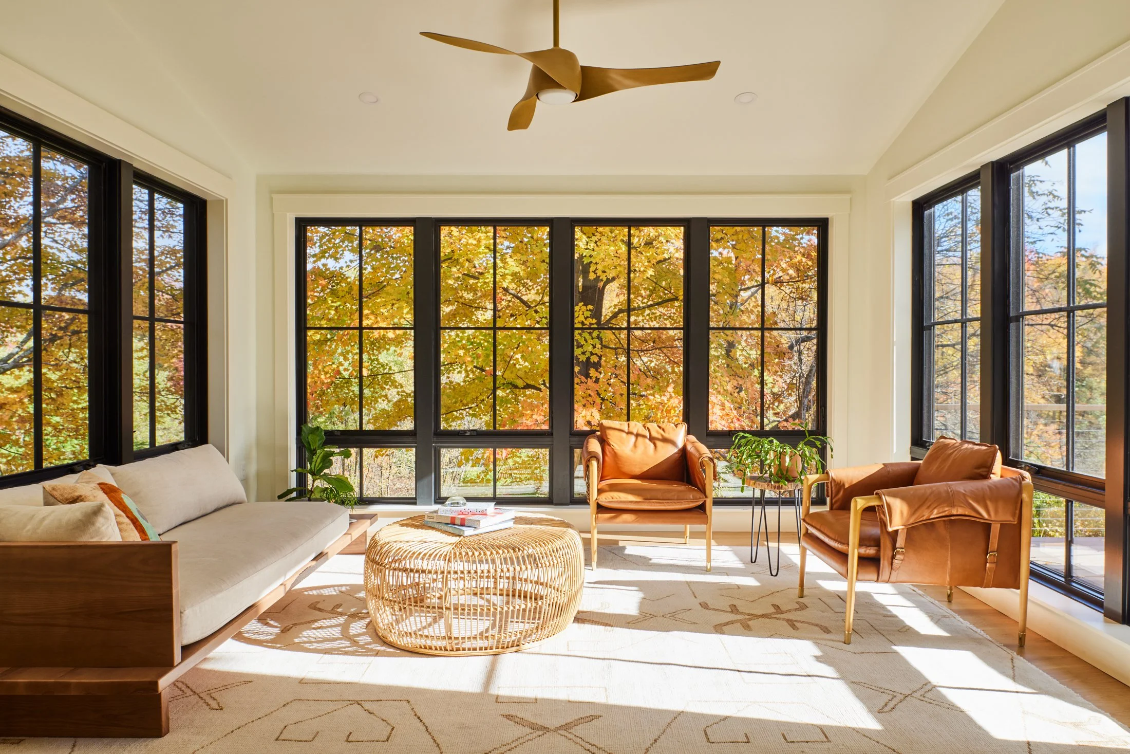 Bright living room with large windows showing fall foliage outside, featuring a beige sofa, two brown leather armchairs, a rattan coffee table, green plants, and a ceiling fan.