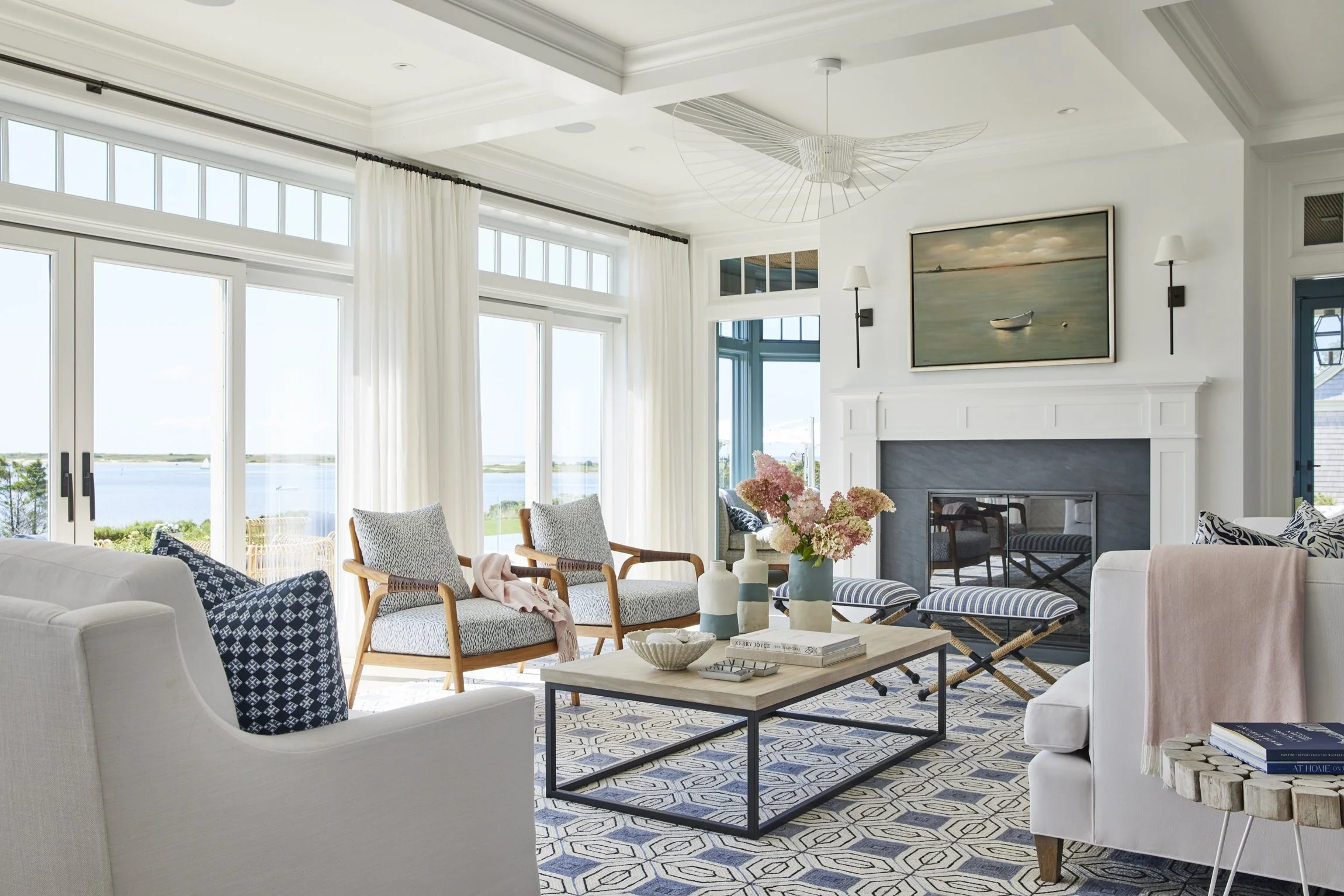 Living room with white walls, a fireplace with a painting above it, large glass doors with white curtains, and coastal decor including pink flowers on a wooden coffee table.