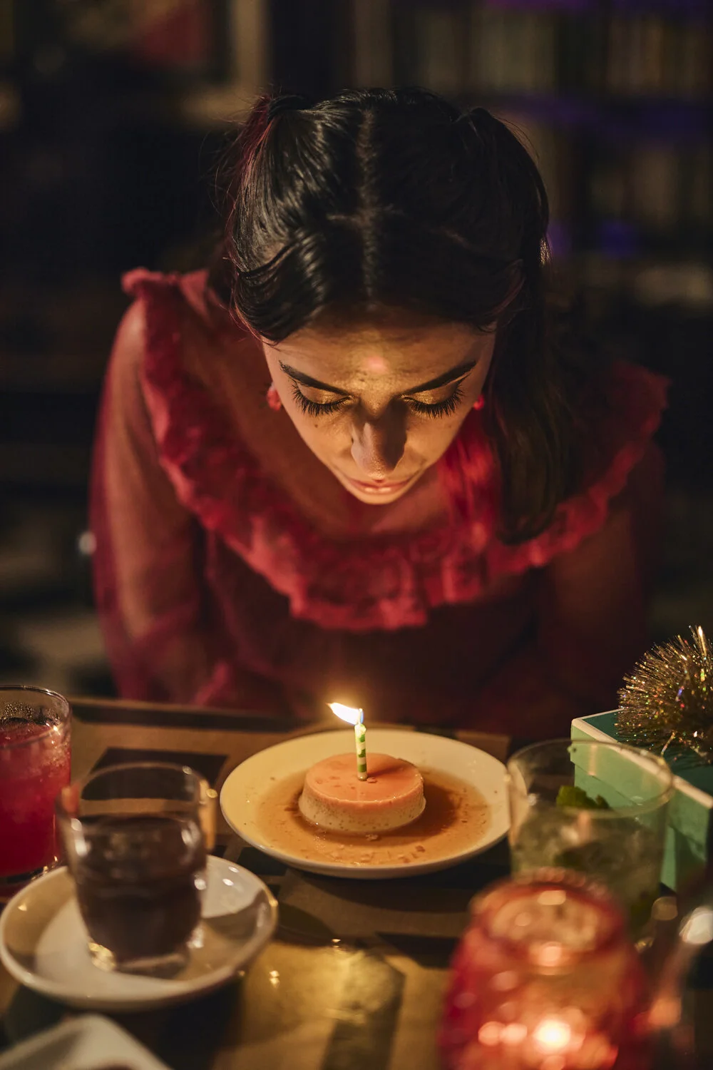 A woman with dark hair leaning over a birthday dessert with a lit candle on top, surrounded by drinks and festive decorations.