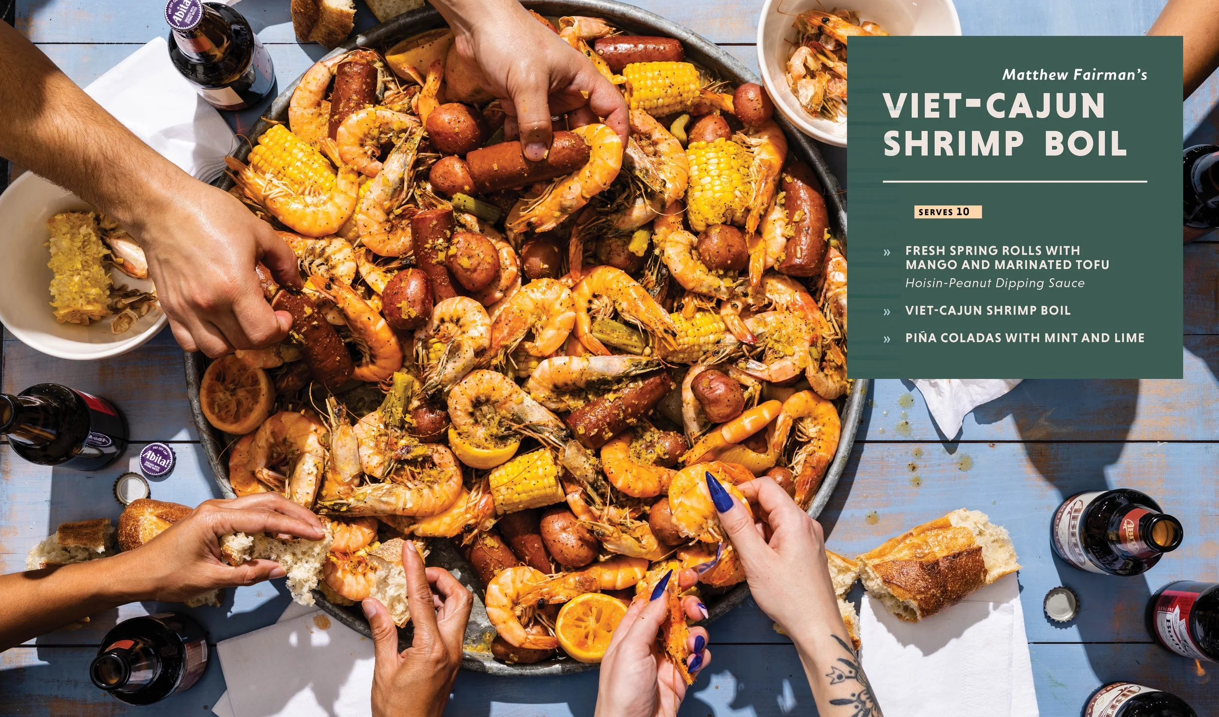 A large tray of cooked shrimp and sausage with corn on the cob on a blue wooden table, surrounded by bottles of beer, bread, and small bowls of dipping sauce, with hands reaching in to serve food.