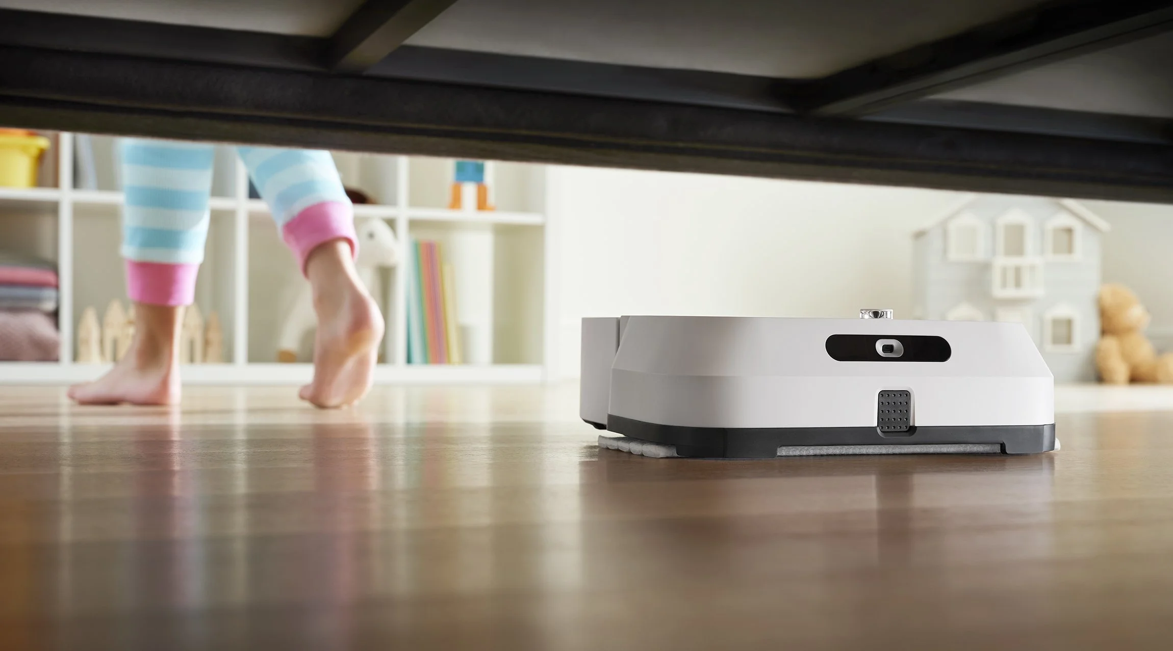 A robotic vacuum cleaner on a wooden floor in a child's room with a child in pajamas visible from the knees down. Photographed by Joseph Keller for iRobot