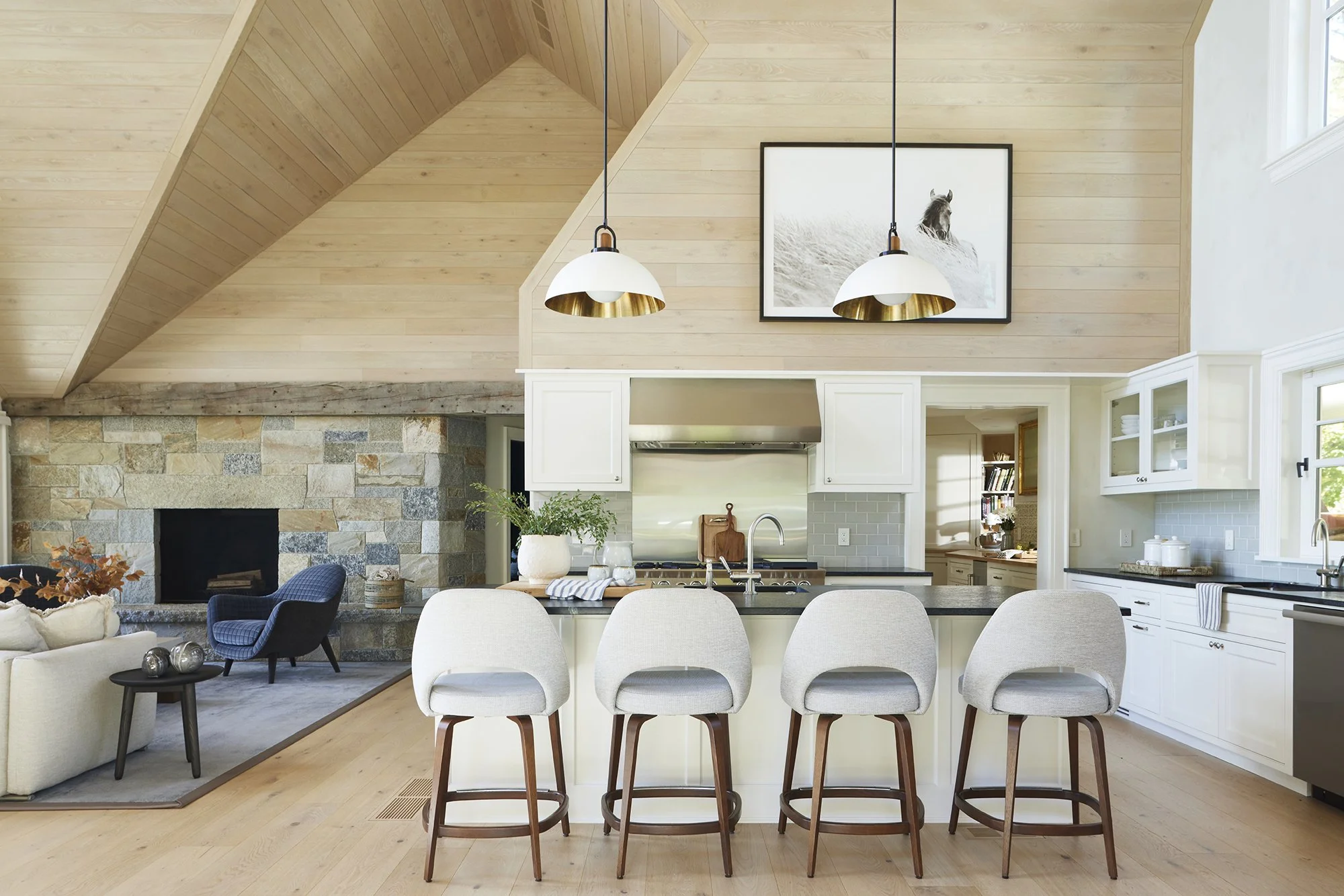 Open kitchen with four barstools, white cabinets, granite counter, and a stone fireplace in a living room with wood-paneled sloped ceiling and framed horse photograph.