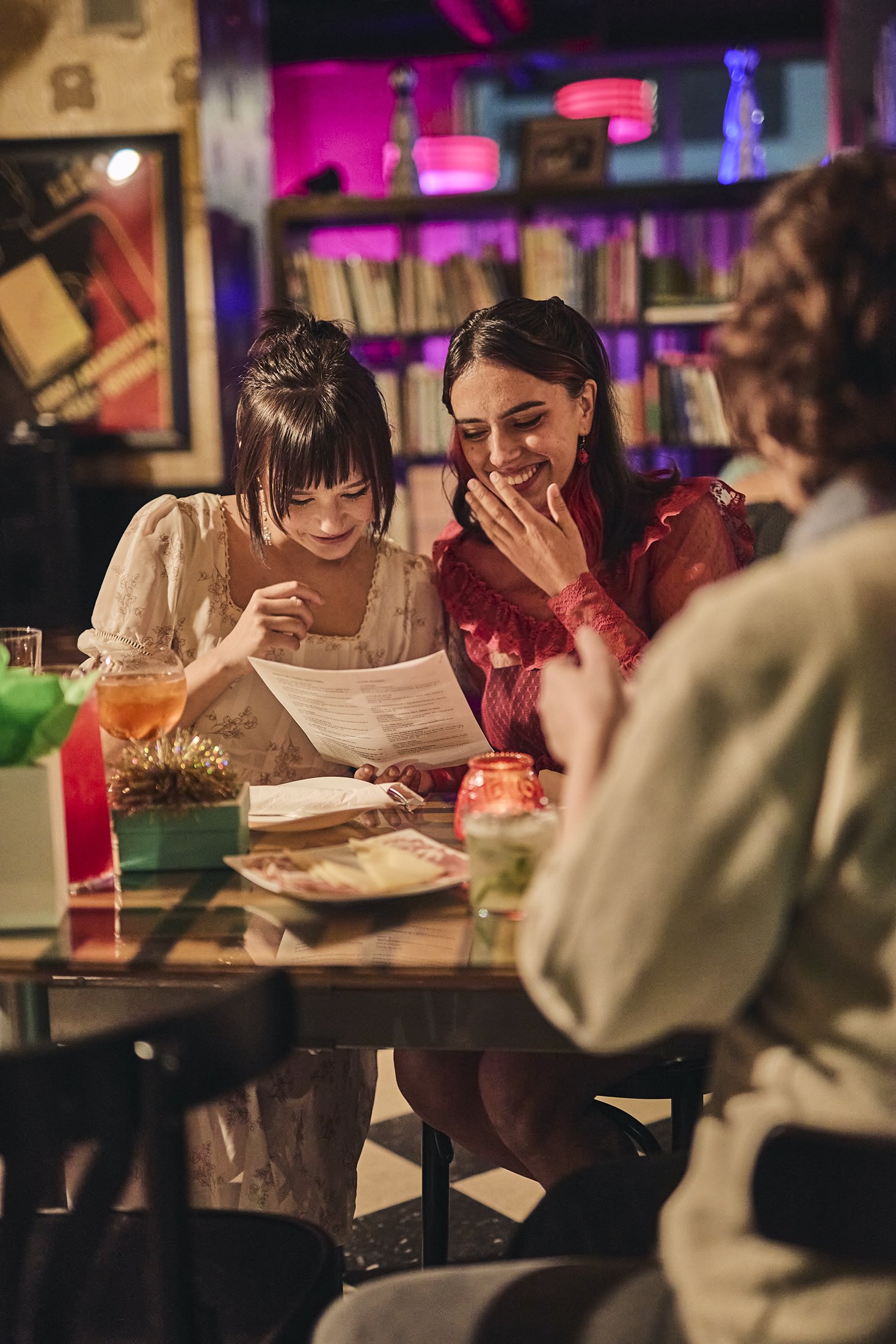 Three women sitting at a table in a cozy, dimly lit bar or restaurant, looking at a menu and laughing, decorated for the holidays with wrapped gifts, candles, and festive decor.