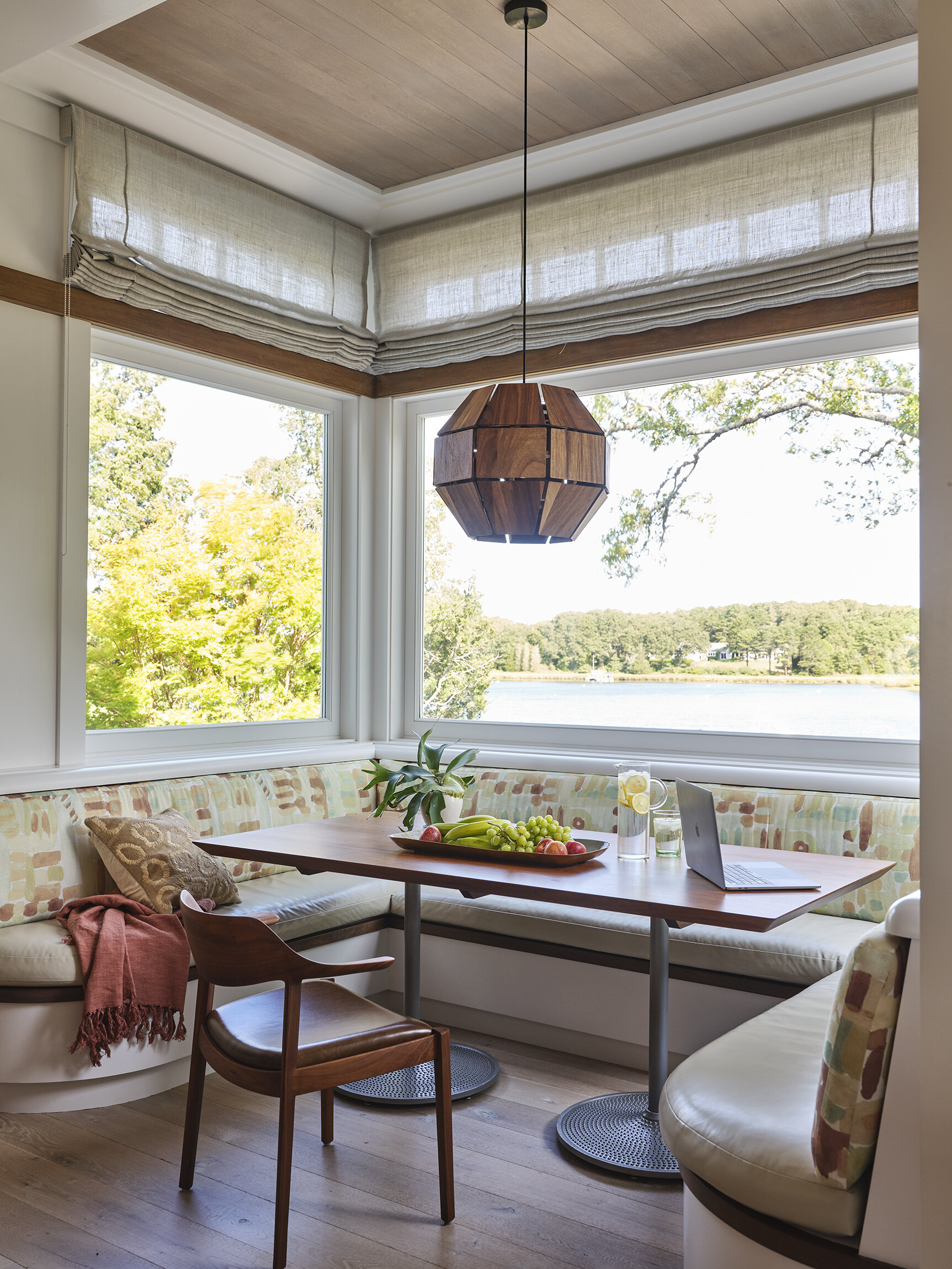 Sunlit dining nook with curved seating, a wooden table, a laptop, fruit bowl, glass of water, and large windows overlooking a river and wooded landscape.