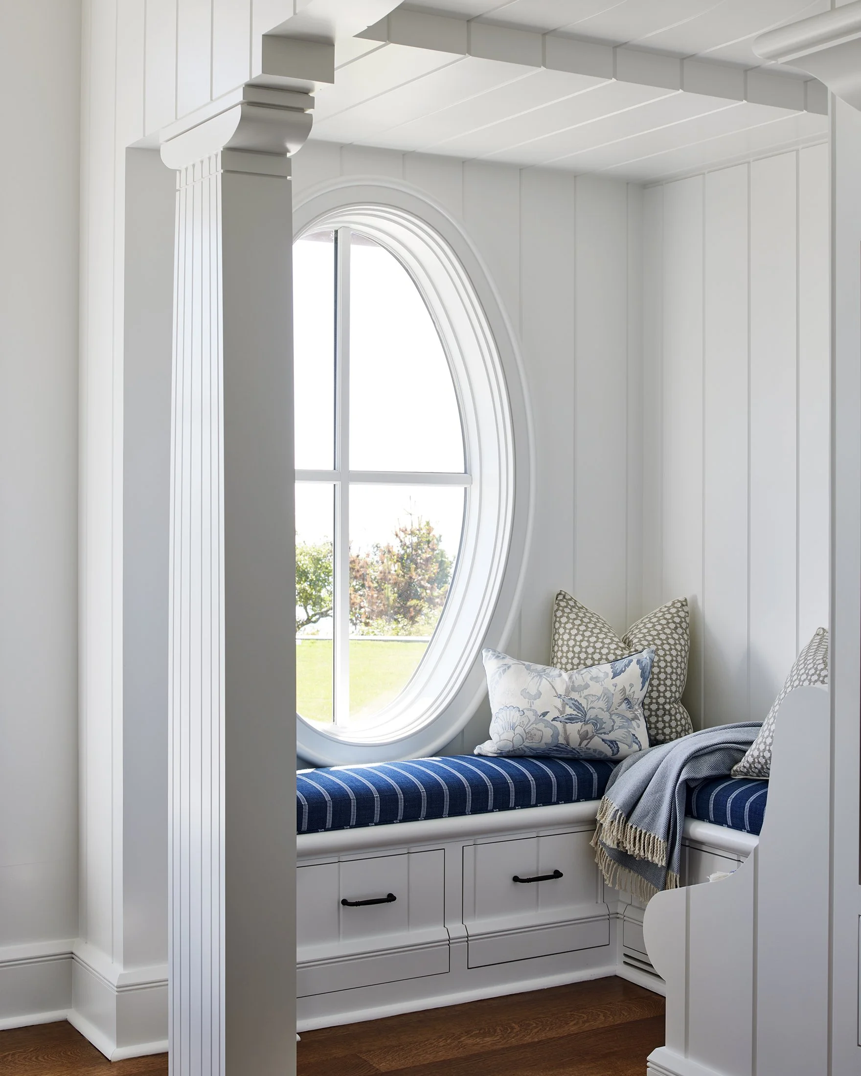 White window seat with blue striped cushion and patterned pillows beneath an oval window in a bright white room with wood flooring.