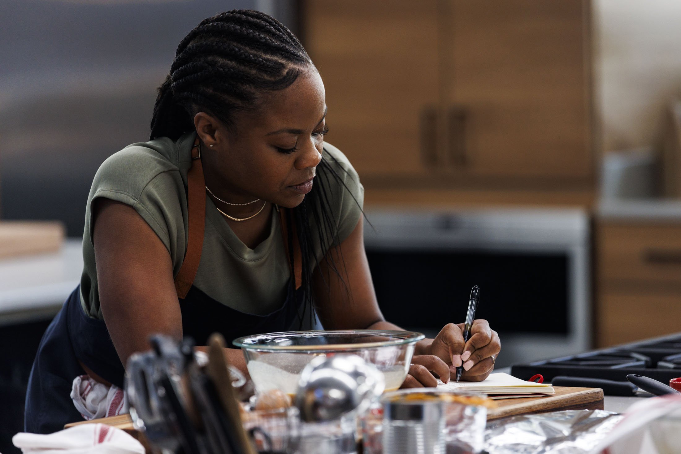 A woman with braided hair writing in a notebook in a kitchen, surrounded by cooking utensils and ingredients.