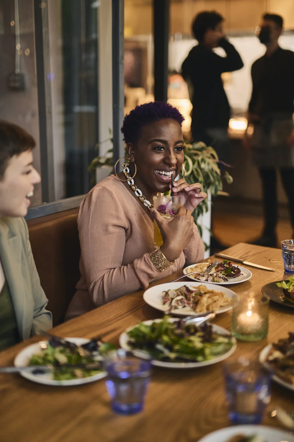 A woman with short purple hair, wearing large hoop earrings and a beige top, enjoying a cocktail at a dinner table with friends. The table has various dishes and glasses, with a lively restaurant background.