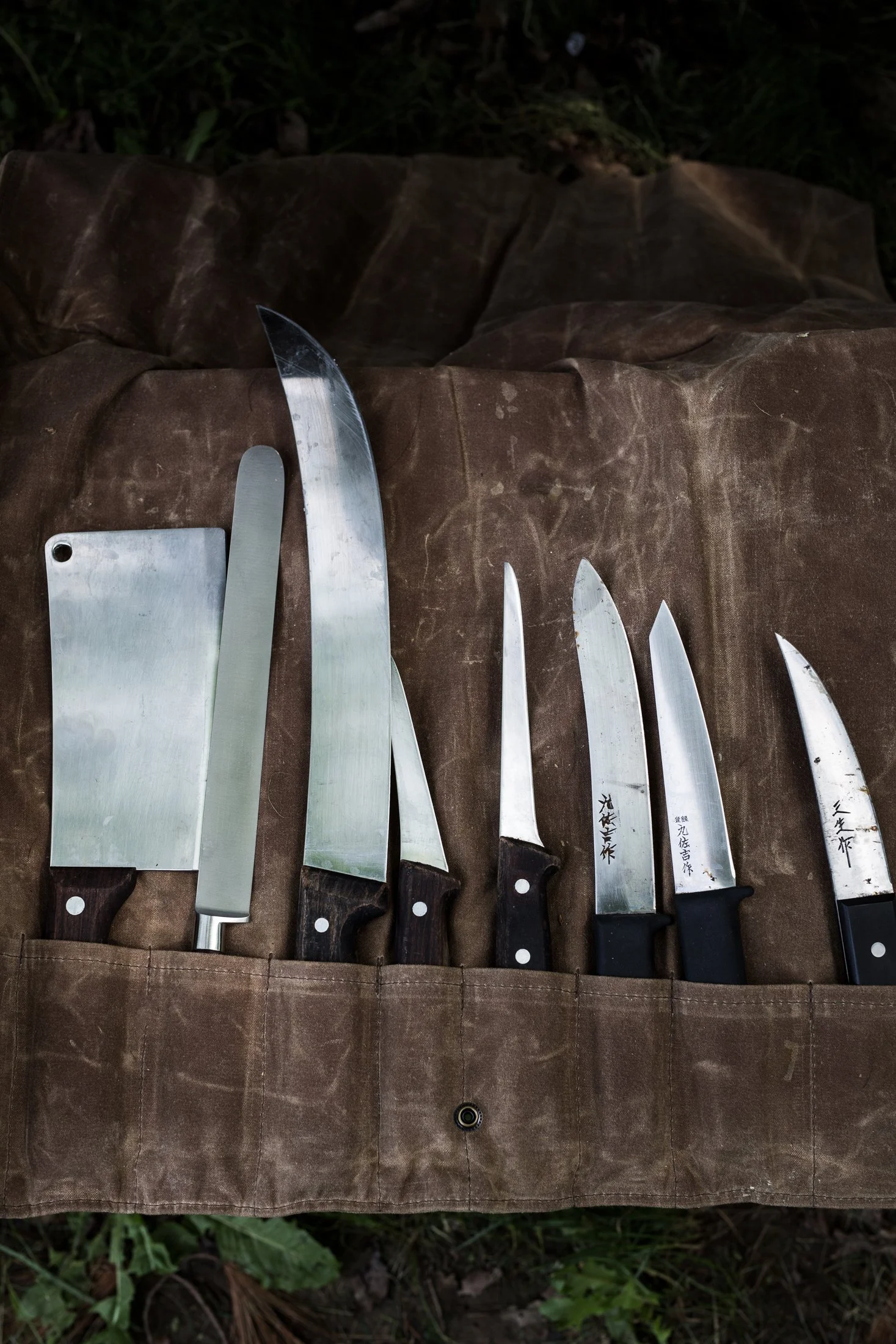 A set of kitchen knives with black handles and Japanese characters on the blades, laid out on a brown fabric surface outdoors. Photographed for the cookbook titled The Smokehouse Handbook