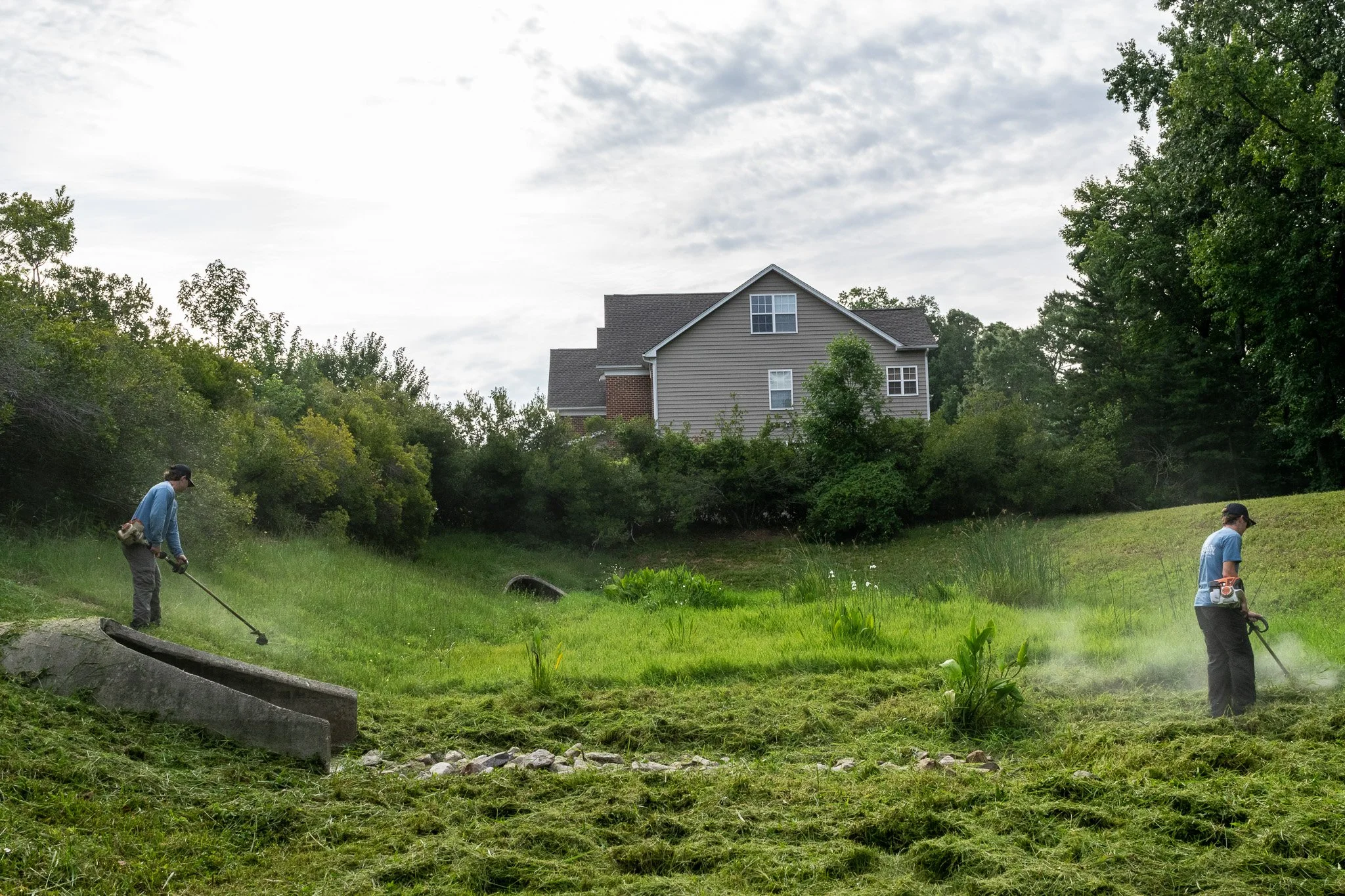 Two stormwater management technicians weed eat the basin and slopes of an HOA dry pond as part of a routine mowing service.