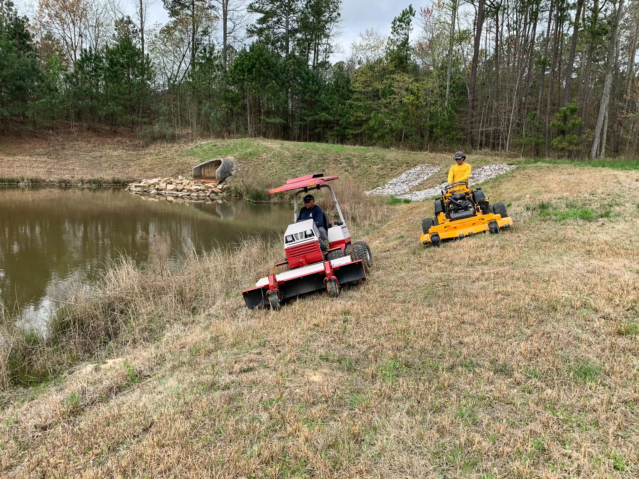 Two stormwater maintenance technicians operate specialized mowing equipment to safely and efficiently mow a retention pond perimeter.