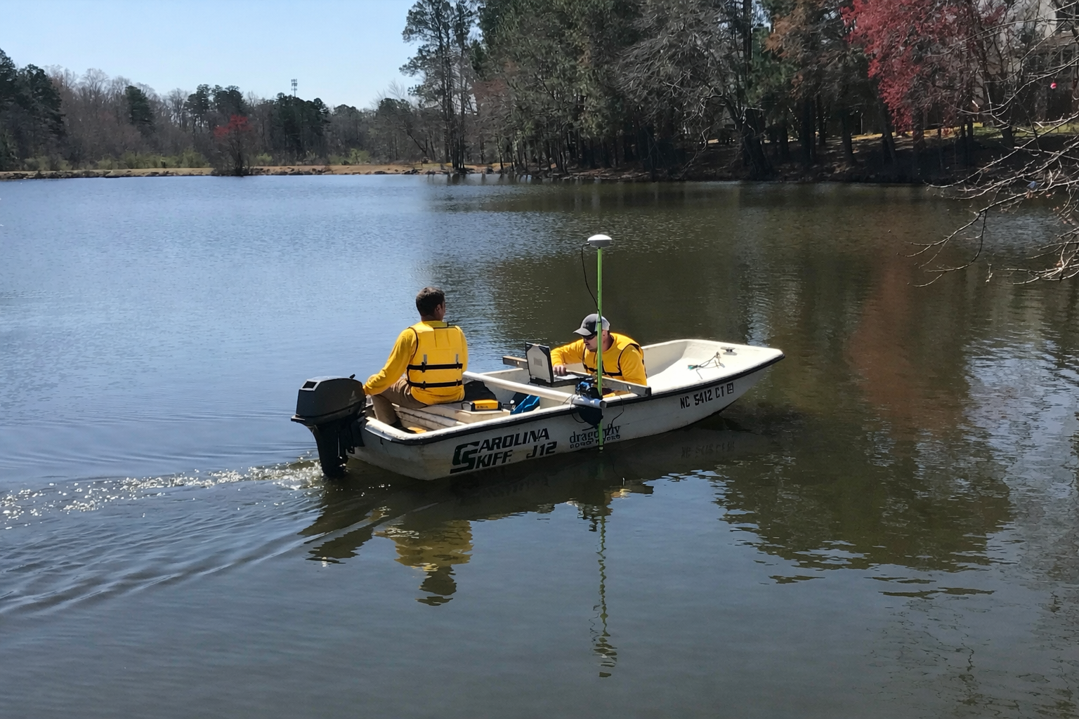 Dragonfly Technicians operate a boat across a calm HOA lake while conducting a bathymetry sediment study to assess sediment accumulation.