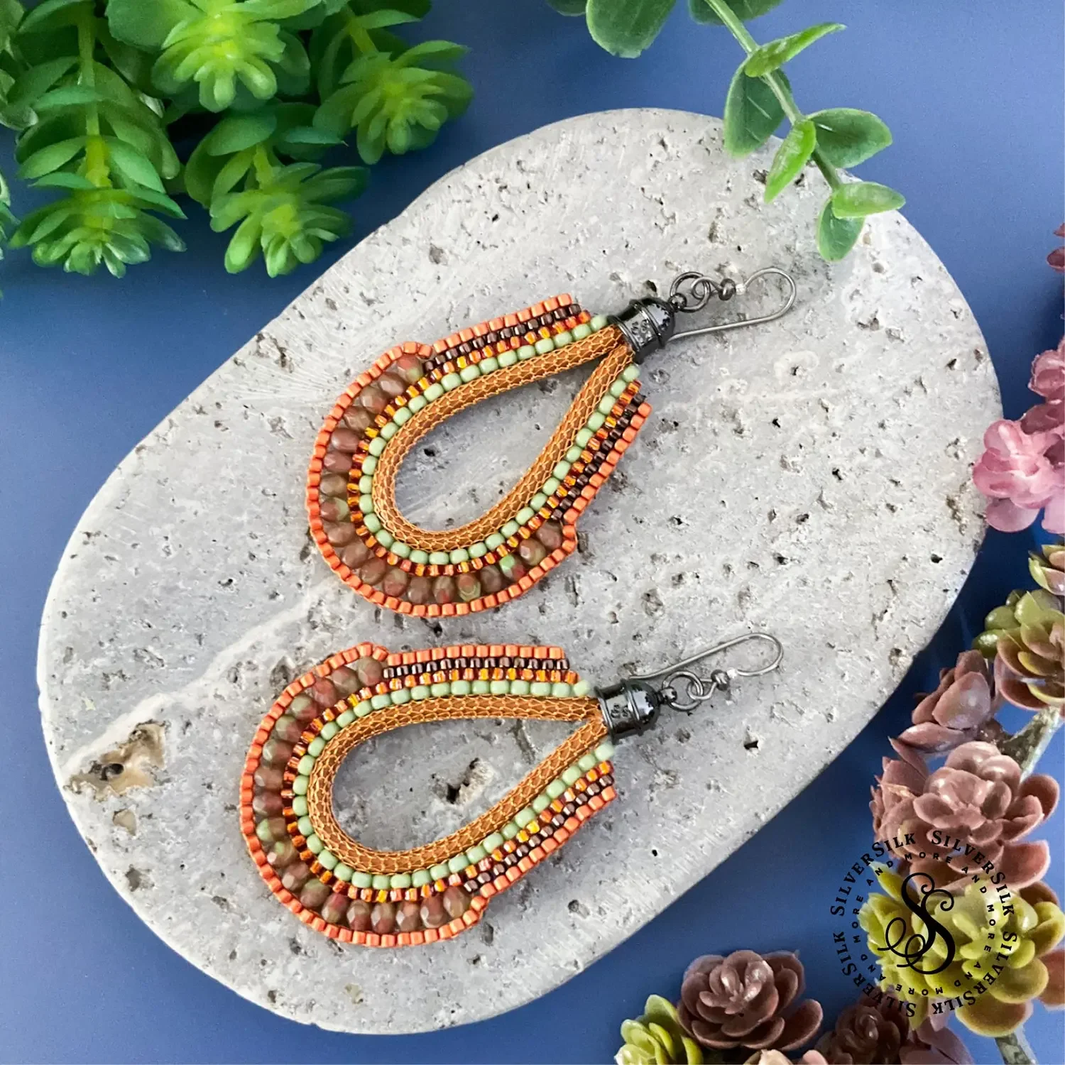 Colorful beaded teardrop-shaped earrings on a textured gray stone with green and purple plants around.