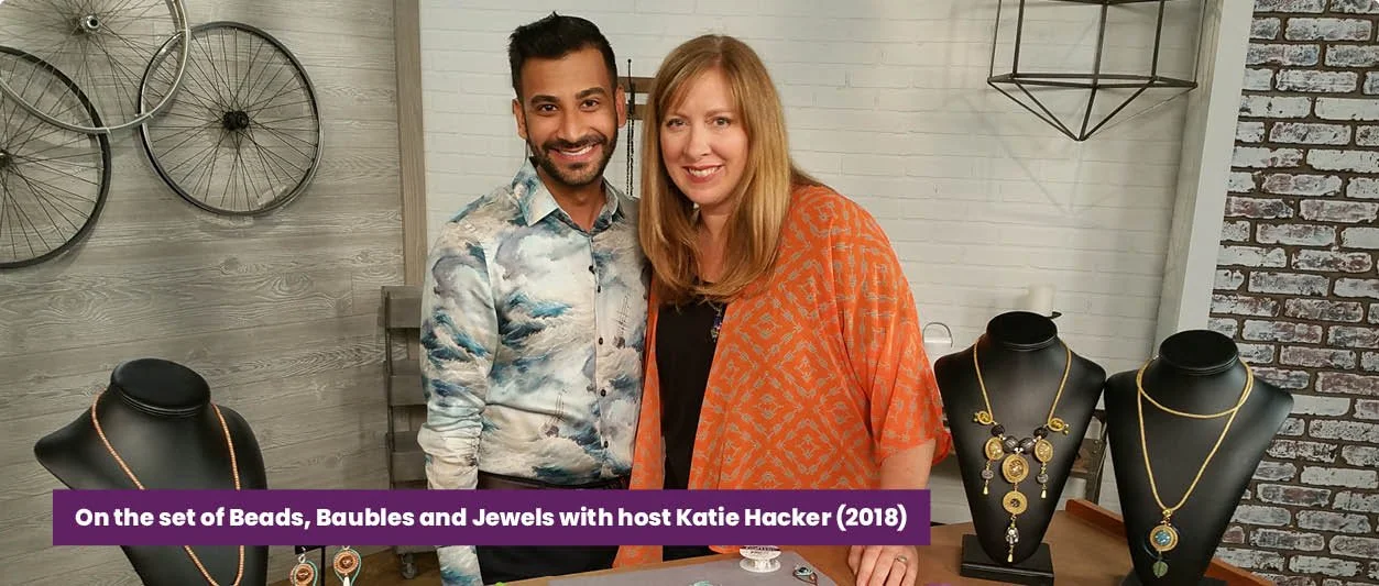 Two people, a man and a woman, stand behind a jewelry display with necklaces on black busts, in a room with decorative wheels on the wall. The caption reads "On the set of Beads, Baubles and Jewels with host Katie Hacker (2018)."