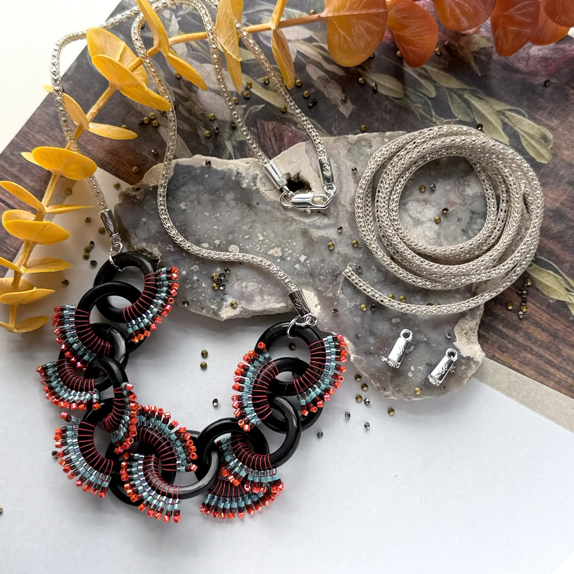 Jewelry displayed on a marble and wooden surface, including a statement necklace with large black and red links and colorful beaded accents, a silver chain necklace, a coiled silver snake chain, and a pair of small silver earrings. Decorative yellow and reddish-brown leaves are partially visible on the side.